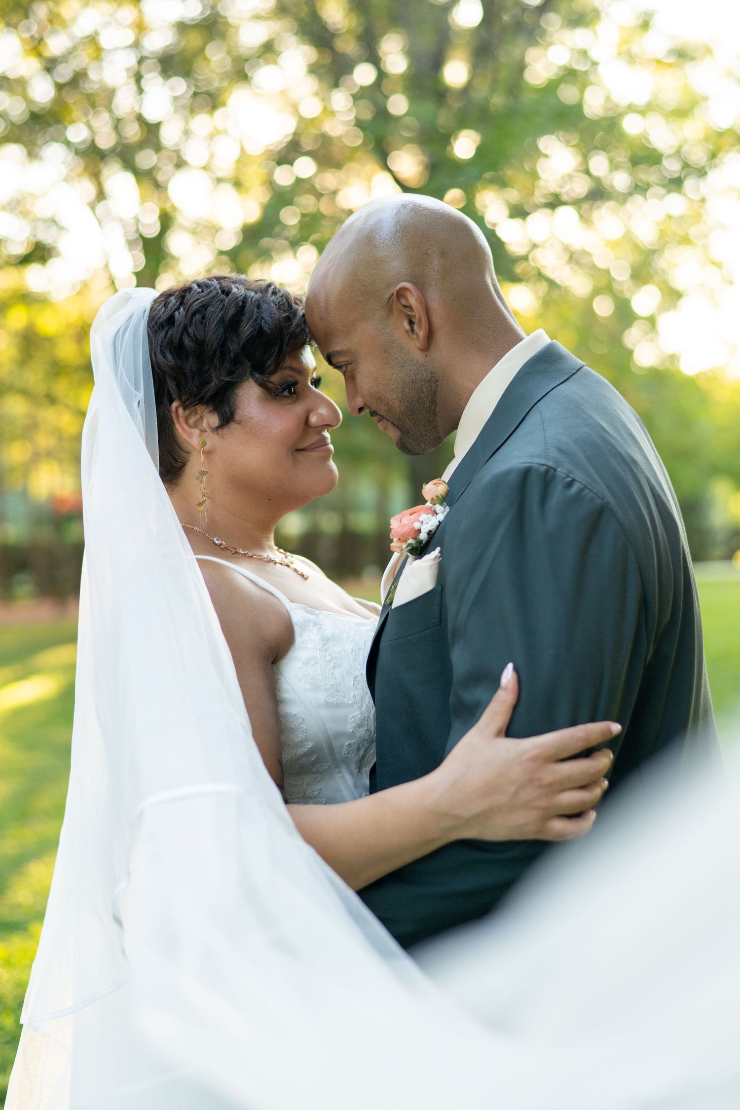 Bride and groom share a romantic forehead touch outdoors, bride in white gown with veil, groom in gray suit.