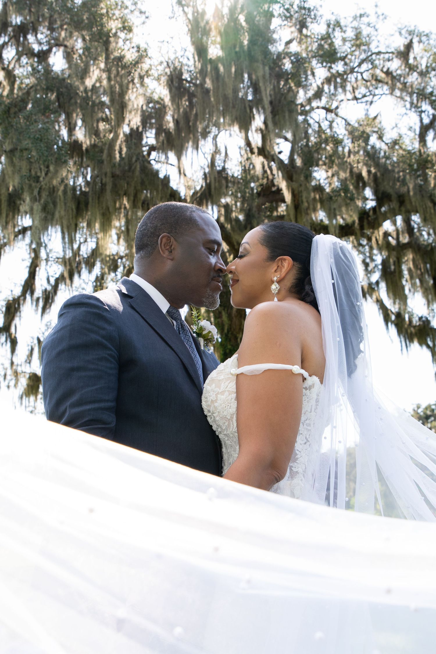 A bride and groom share a romantic moment under moss-draped trees, with her veil flowing gracefully around them.