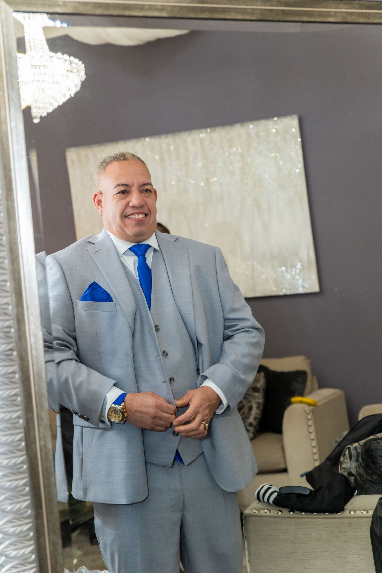 Smiling man in light gray three-piece suit with blue tie and pocket square, buttoning jacket in elegant room.