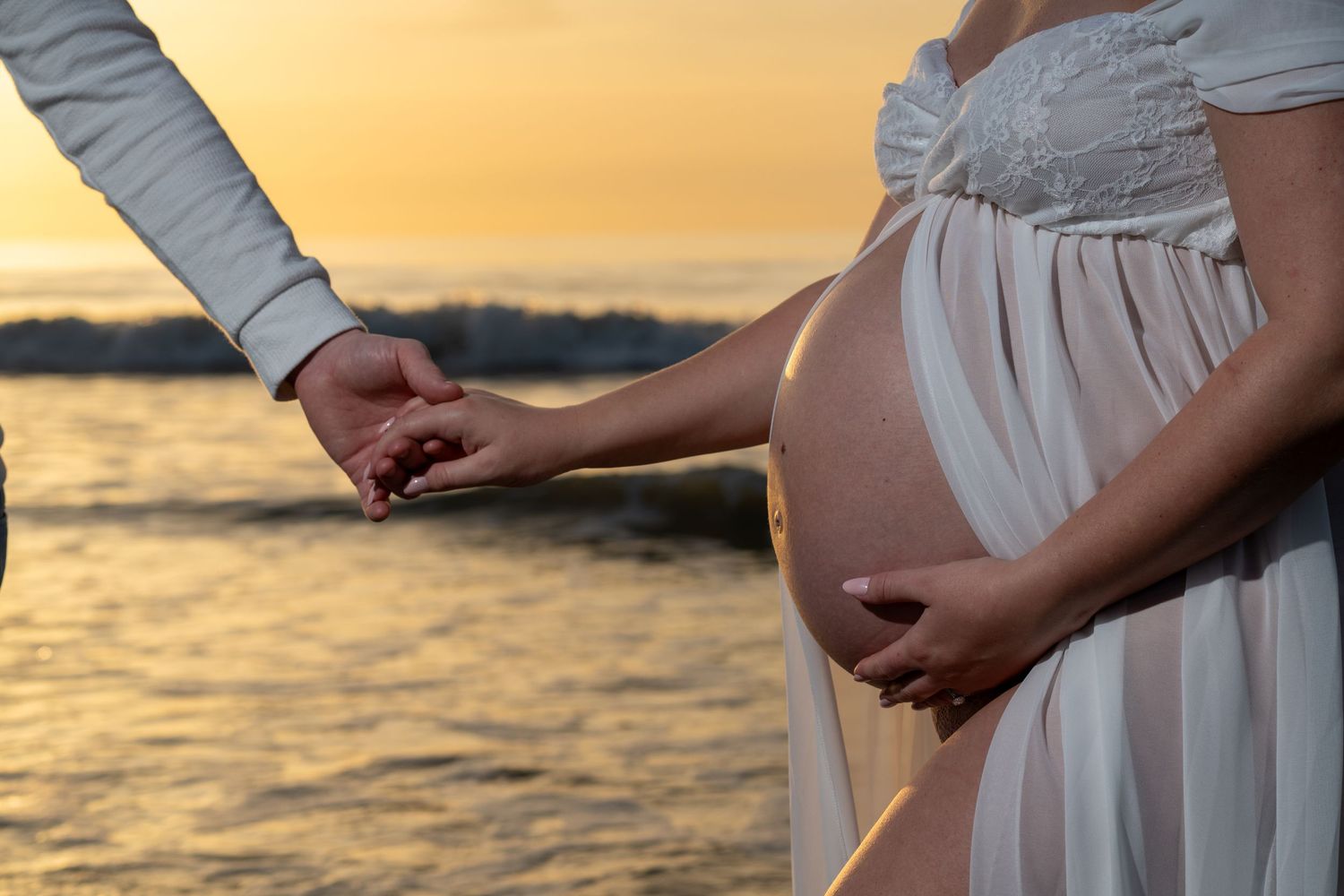Pregnant woman in white dress holds partner's hand by the ocean at golden sunset.