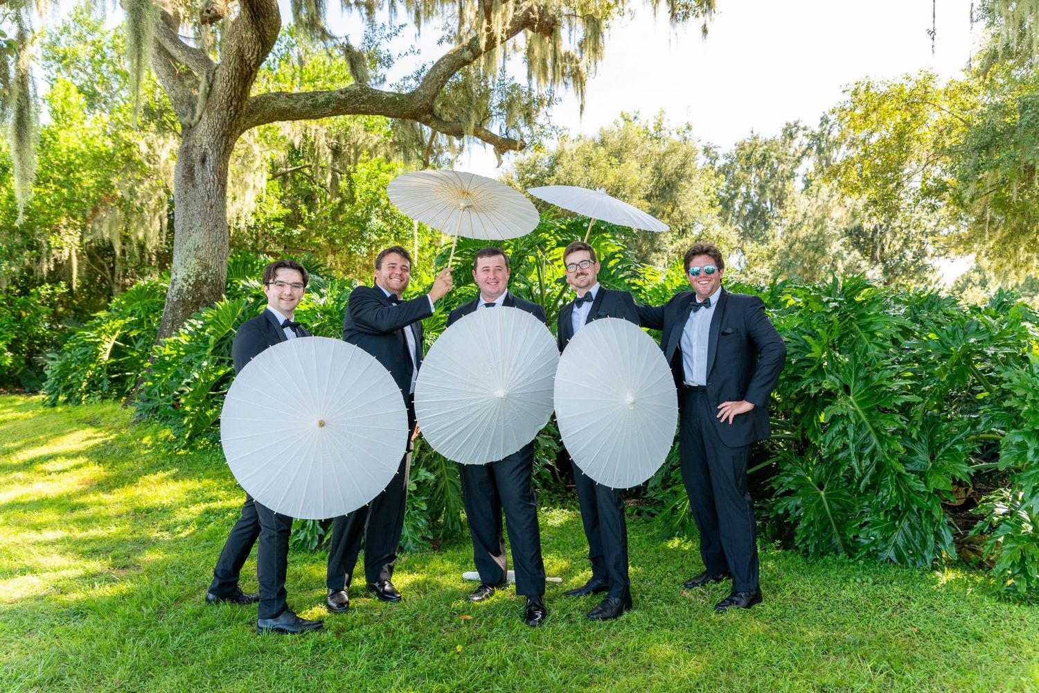 Five groomsmen in tuxedos holding white parasol umbrellas outdoors under a moss-draped oak tree on a sunny day.