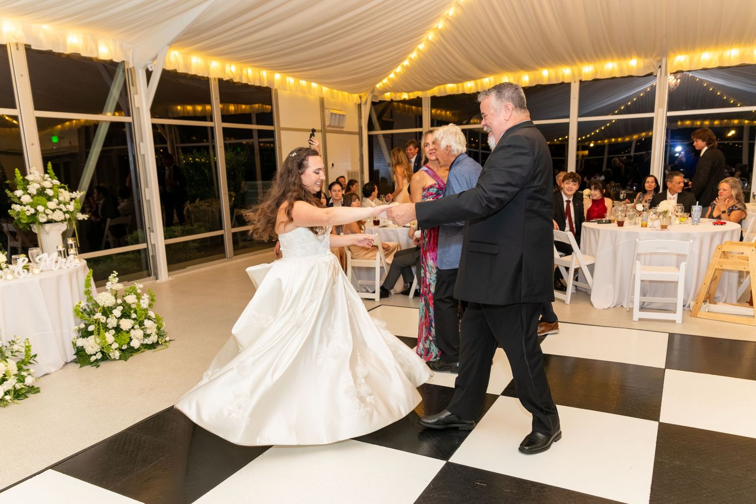 Bride in white gown dancing with a man in a black suit on a checkered dance floor at an elegant wedding reception.