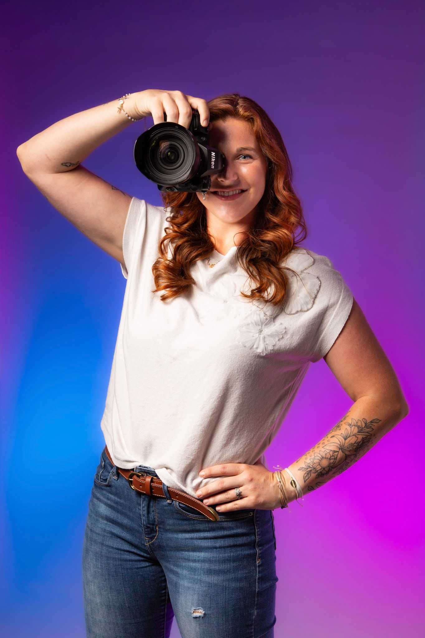 Red-haired female photographer holding DSLR camera, wearing white t-shirt and jeans, posing against colorful purple-blue backdrop.