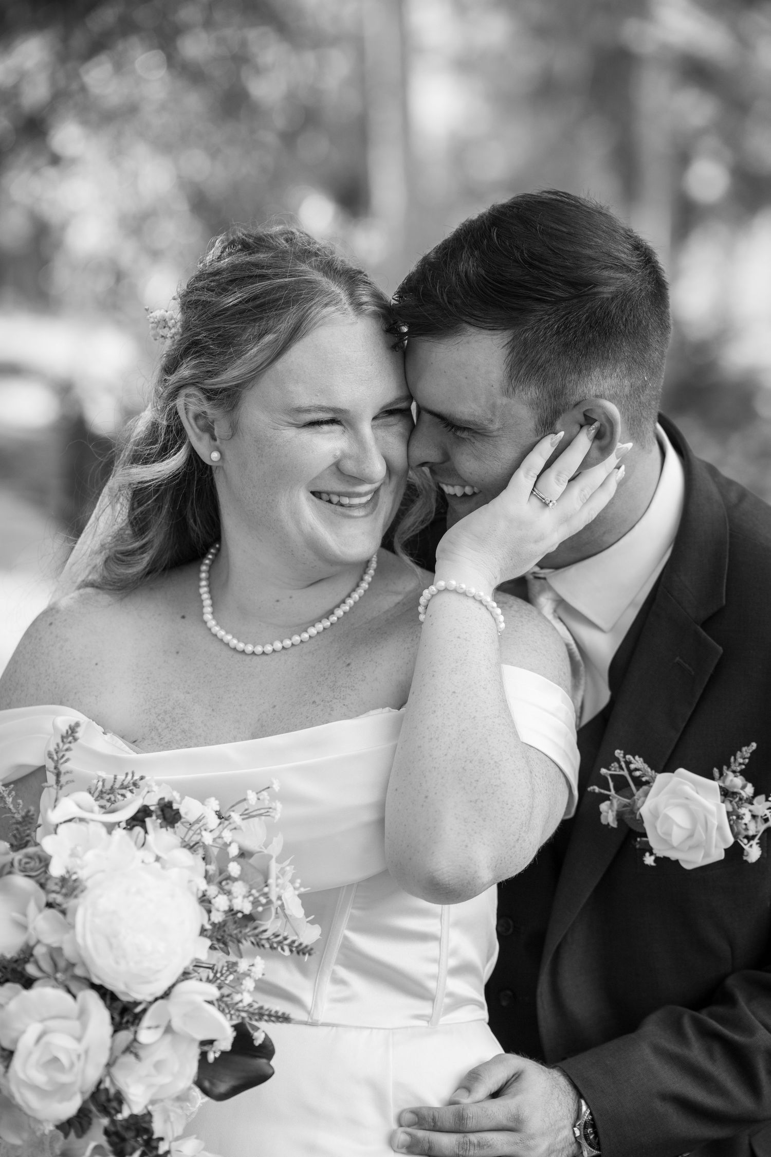 Black and white photo of a smiling bride in strapless gown holding bouquet, leaning close with groom in suit.