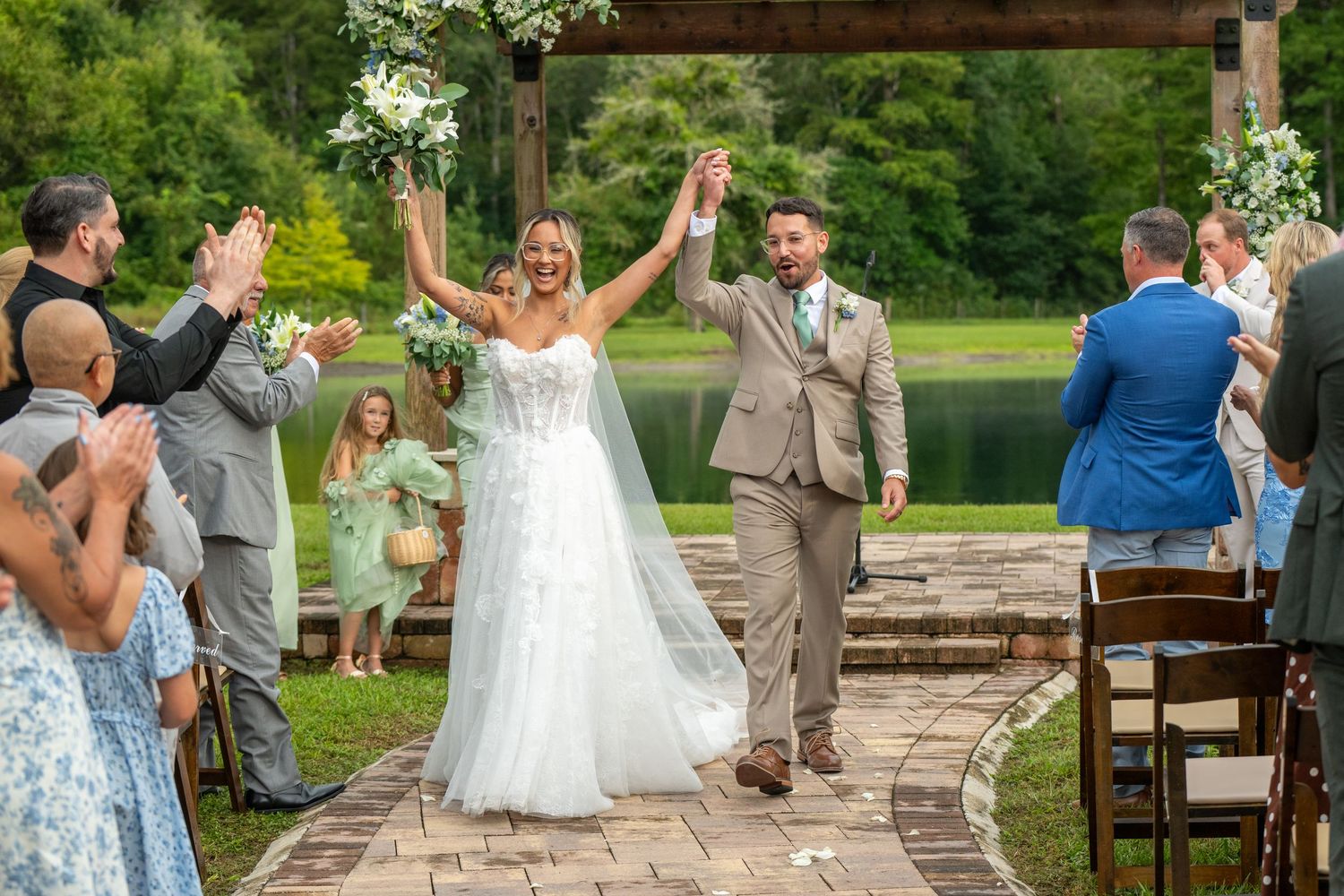 Joyful newlyweds raise hands walking down outdoor aisle surrounded by cheering guests at a lakeside wedding ceremony.