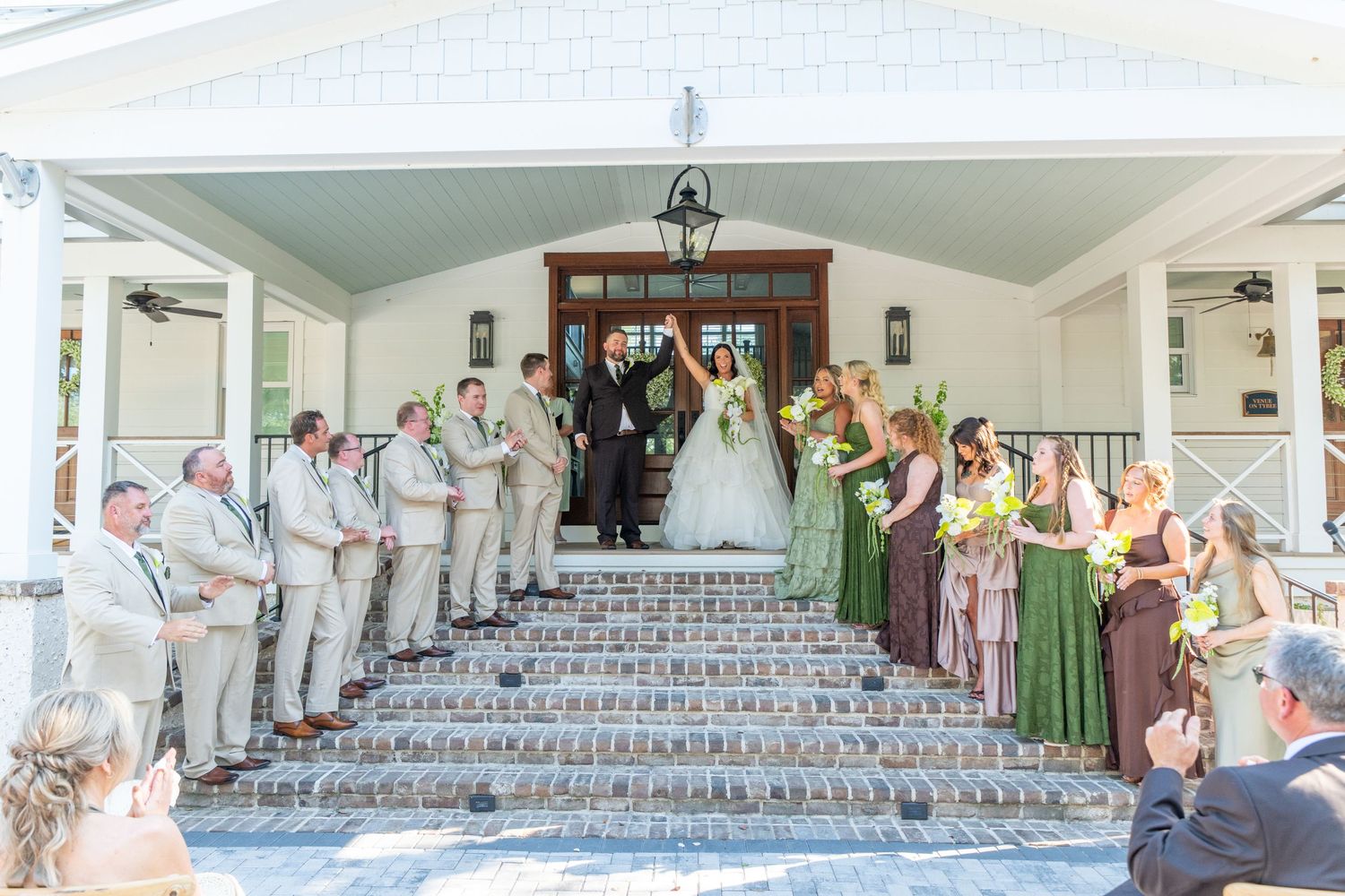 Wedding party poses on brick steps of elegant venue, bride and groom centered with groomsmen and bridesmaids in green dresses.