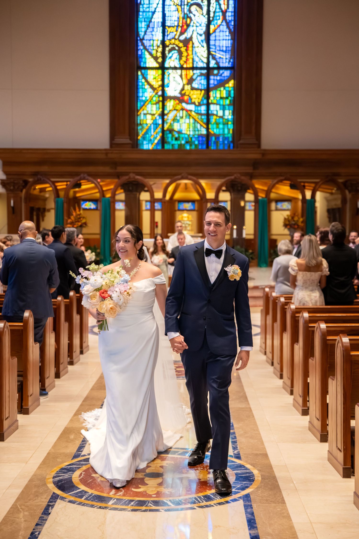 Bride and groom walk down church aisle after wedding ceremony, stained glass window backdrop, bride holds floral bouquet.
