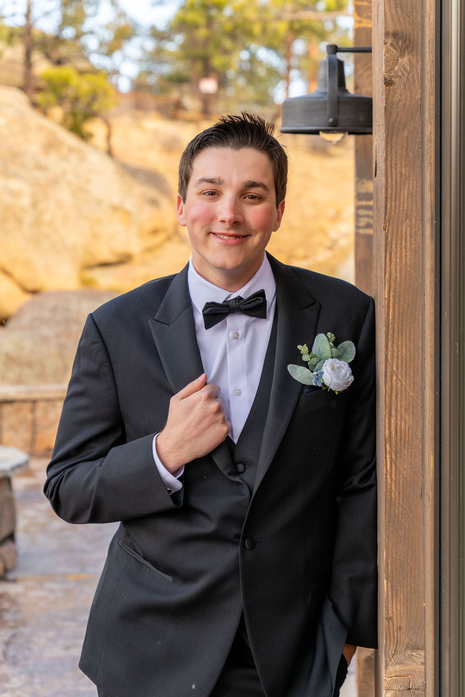 Young groom in black tuxedo with bow tie and white boutonniere smiles while leaning against wooden door outdoors.