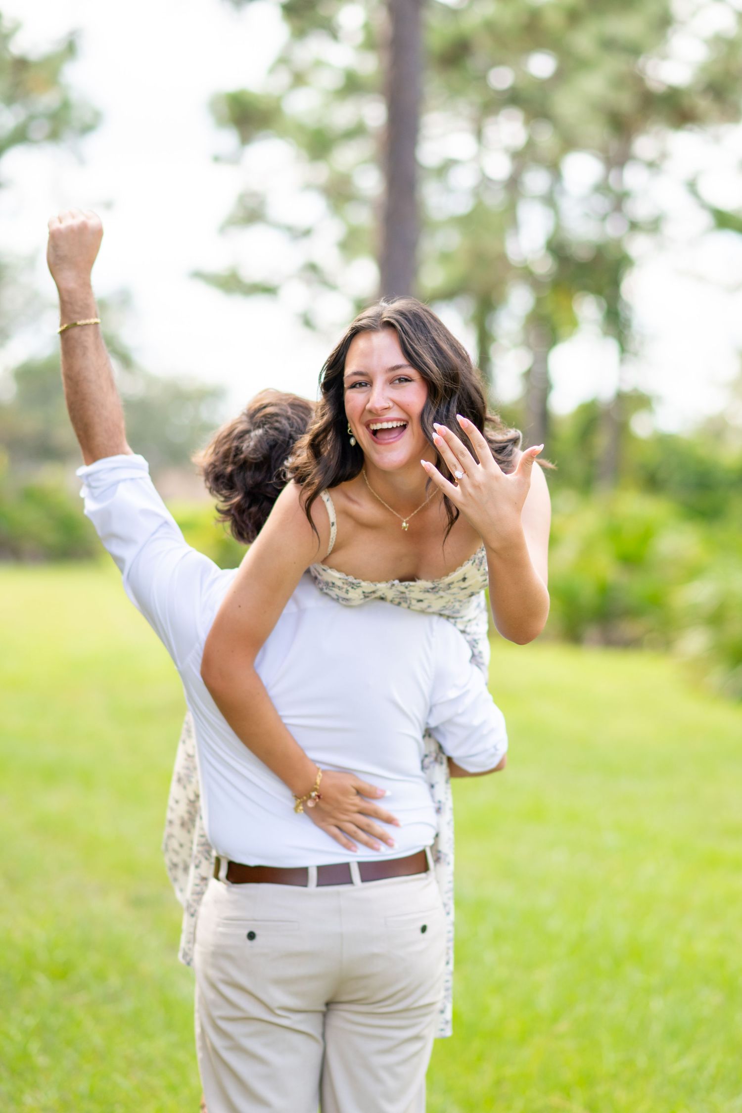 Excited woman shows off engagement ring while her partner lifts her in a sunny park setting.