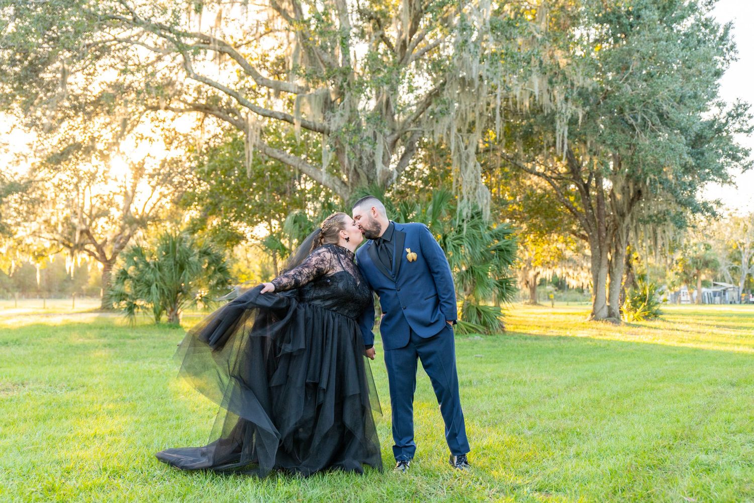 Couple posing romantically in a sunlit park, bride in a flowing black gown and groom in a navy suit.