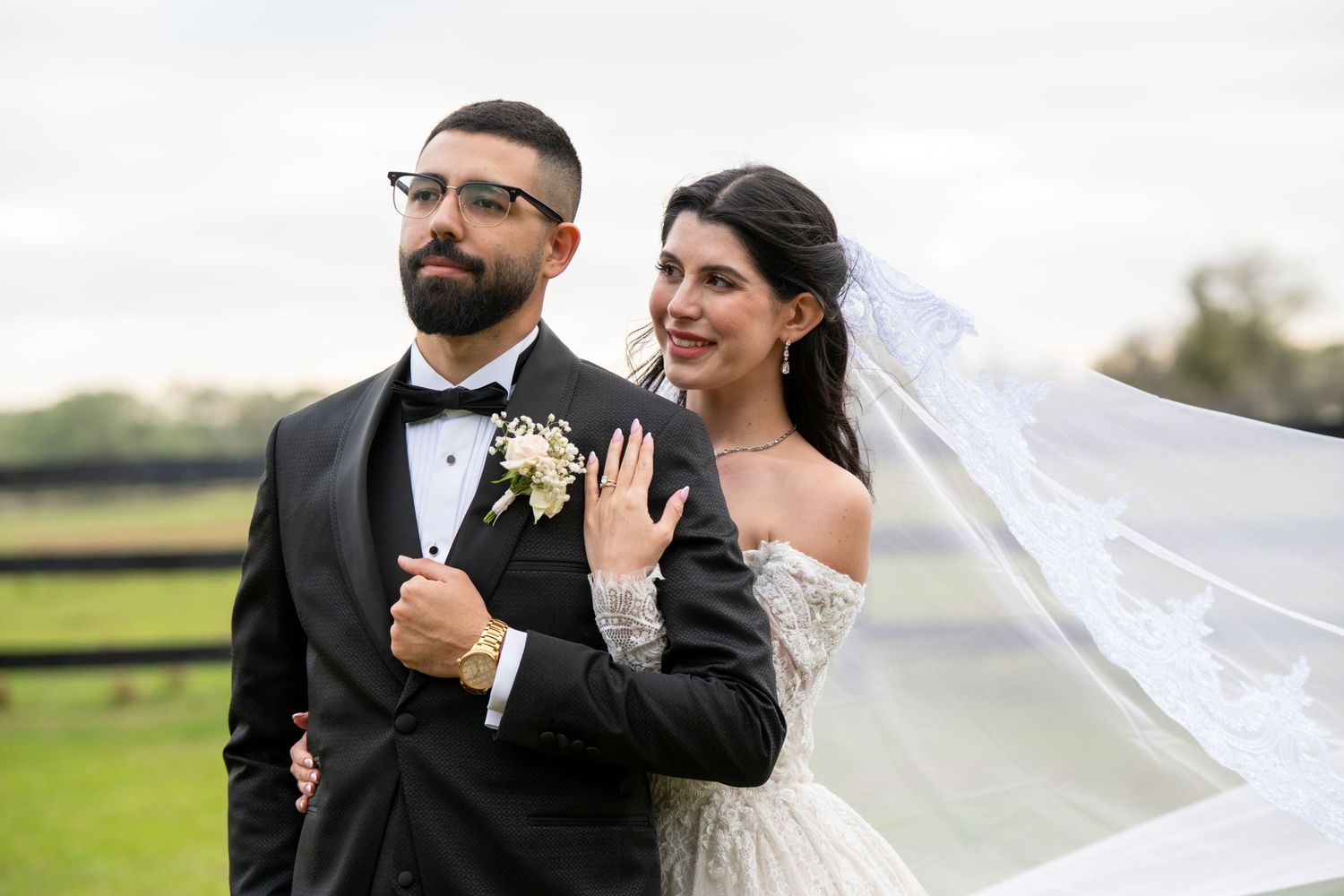 Bride shows off her ring while embracing groom outdoors, her veil flowing in the breeze.