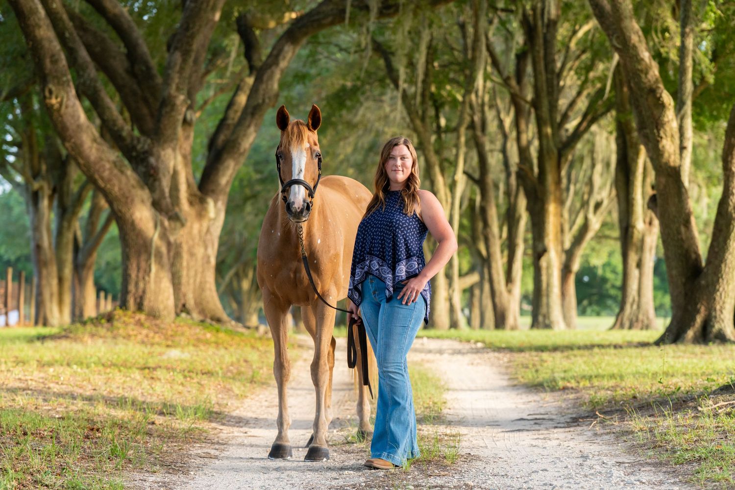 Woman walks chestnut horse down tree-lined dirt path in lush green setting.