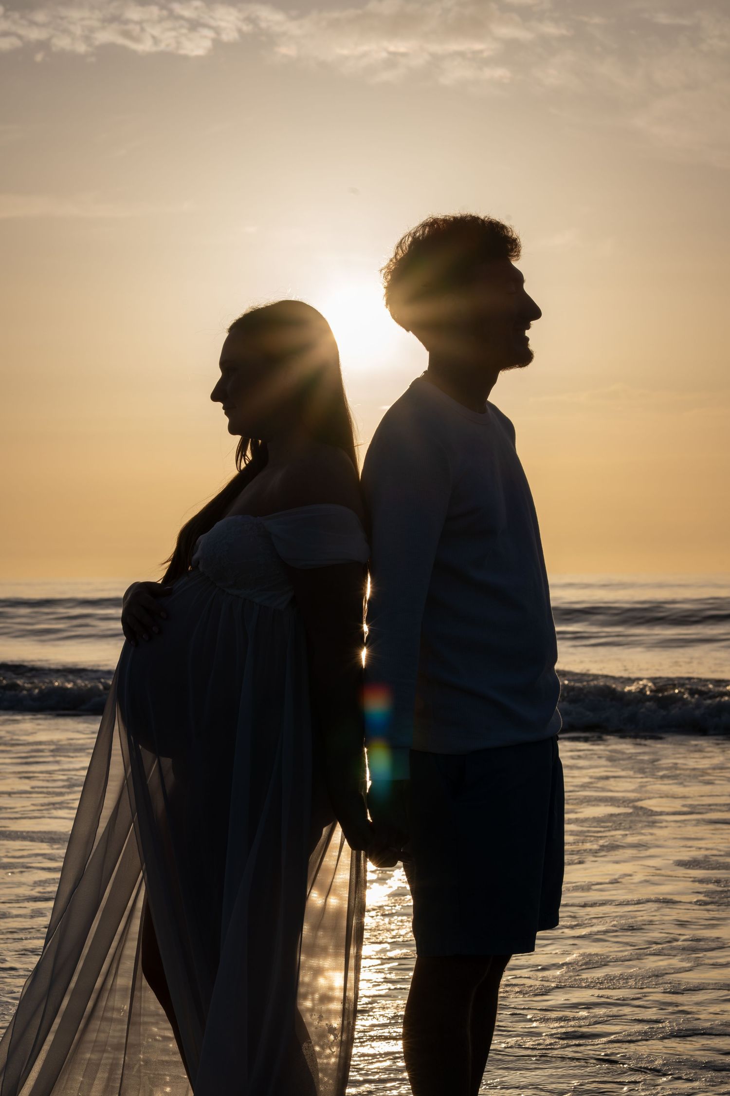 Silhouette of a pregnant couple standing back-to-back on a beach at golden sunset with ocean waves behind them.
