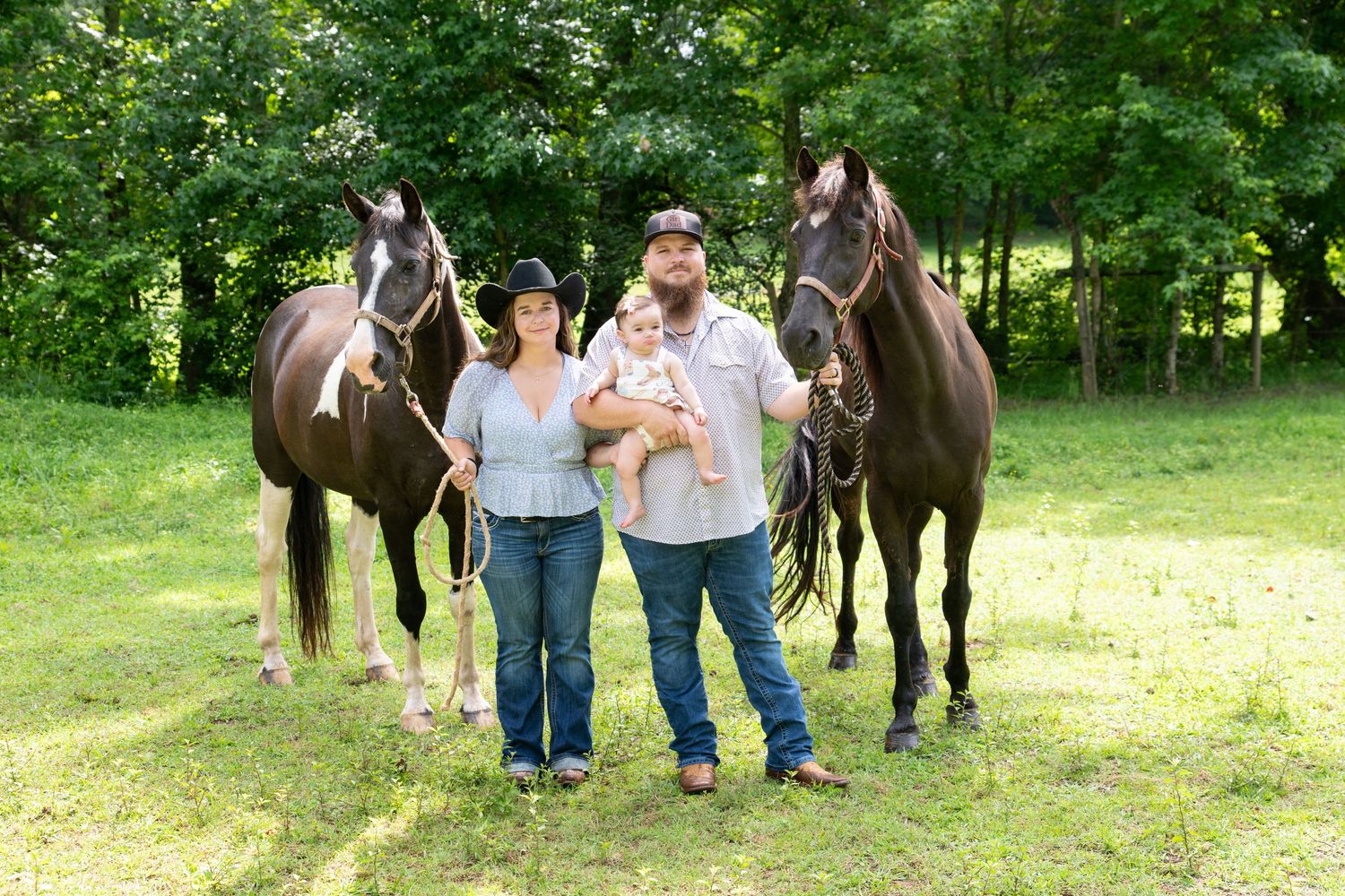 Couple with baby standing between two dark horses on green grass with trees in background.
