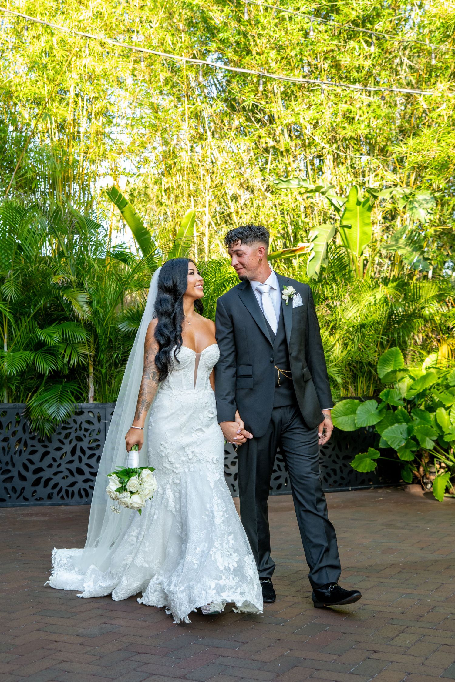 Bride in lace mermaid gown and groom in dark suit hold hands, gazing at each other in a lush tropical garden setting.