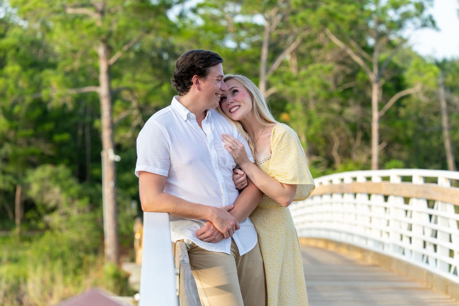 Happy couple embracing on a white bridge surrounded by lush green trees on a sunny day.
