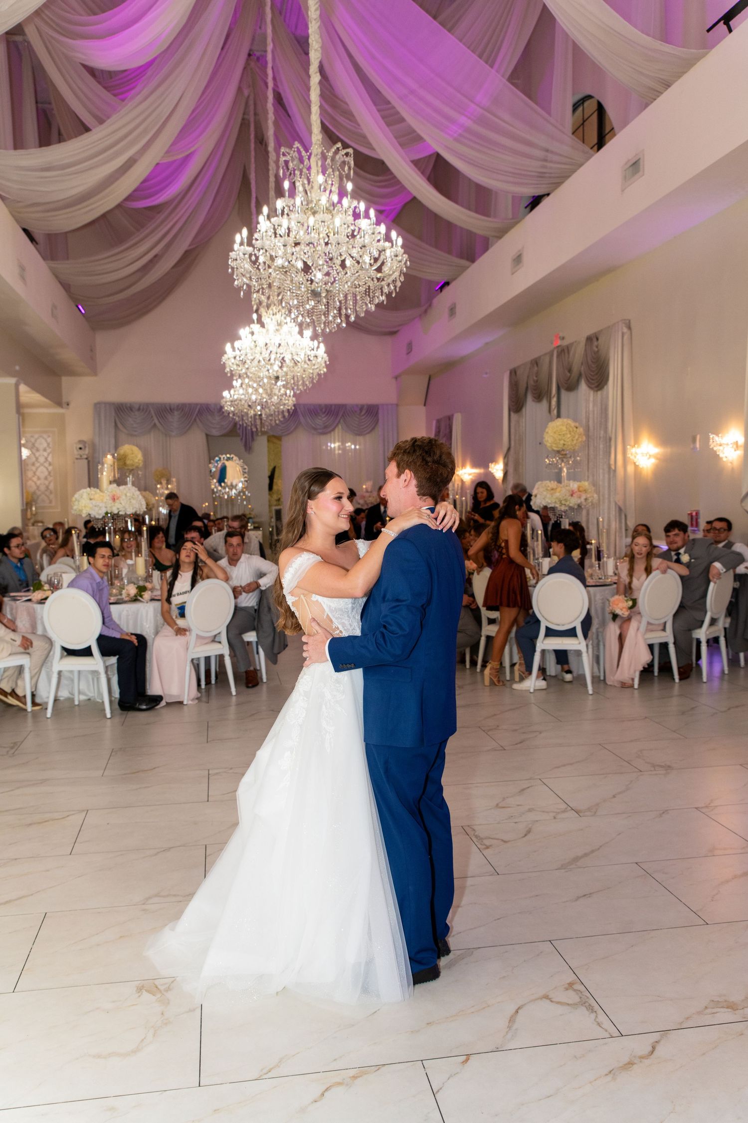 Bride and groom share their first dance under crystal chandeliers in an elegant white ballroom with purple uplighting.