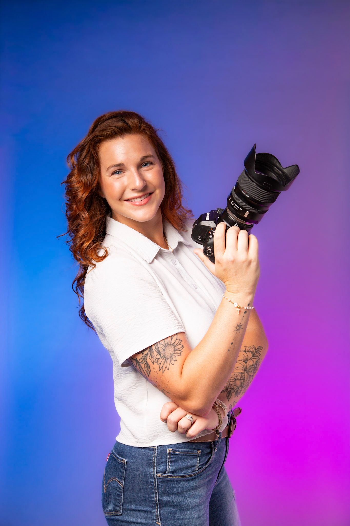 Smiling female photographer holding a DSLR camera, wearing a white shirt and jeans, with tattoos, against a colorful backdrop.