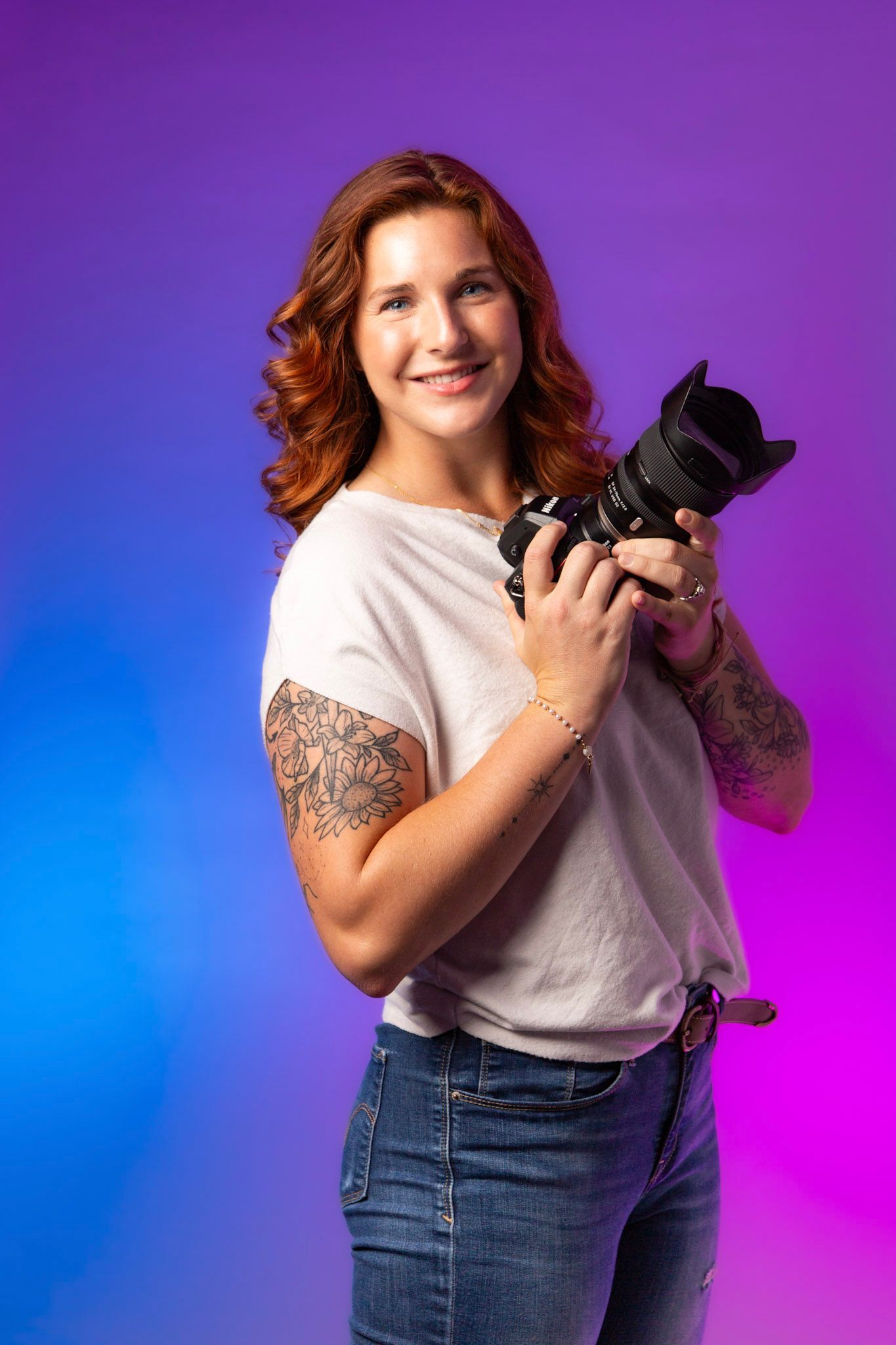 A smiling redhead woman with tattoos holds a DSLR camera, wearing a white tee and jeans against a purple-blue backdrop.