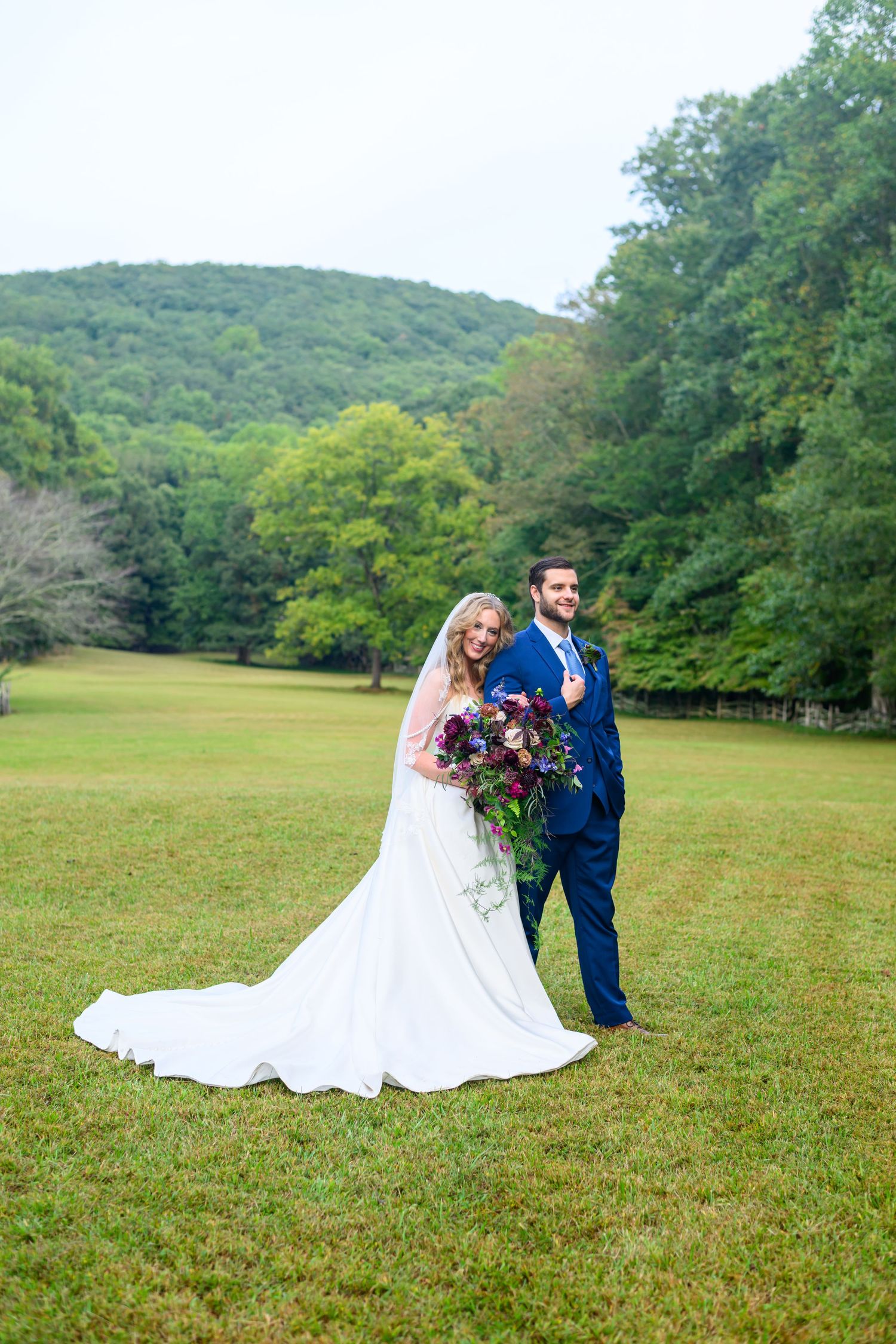 Bride and groom posing on green lawn with lush forested hills, bride holding purple bouquet in white gown.