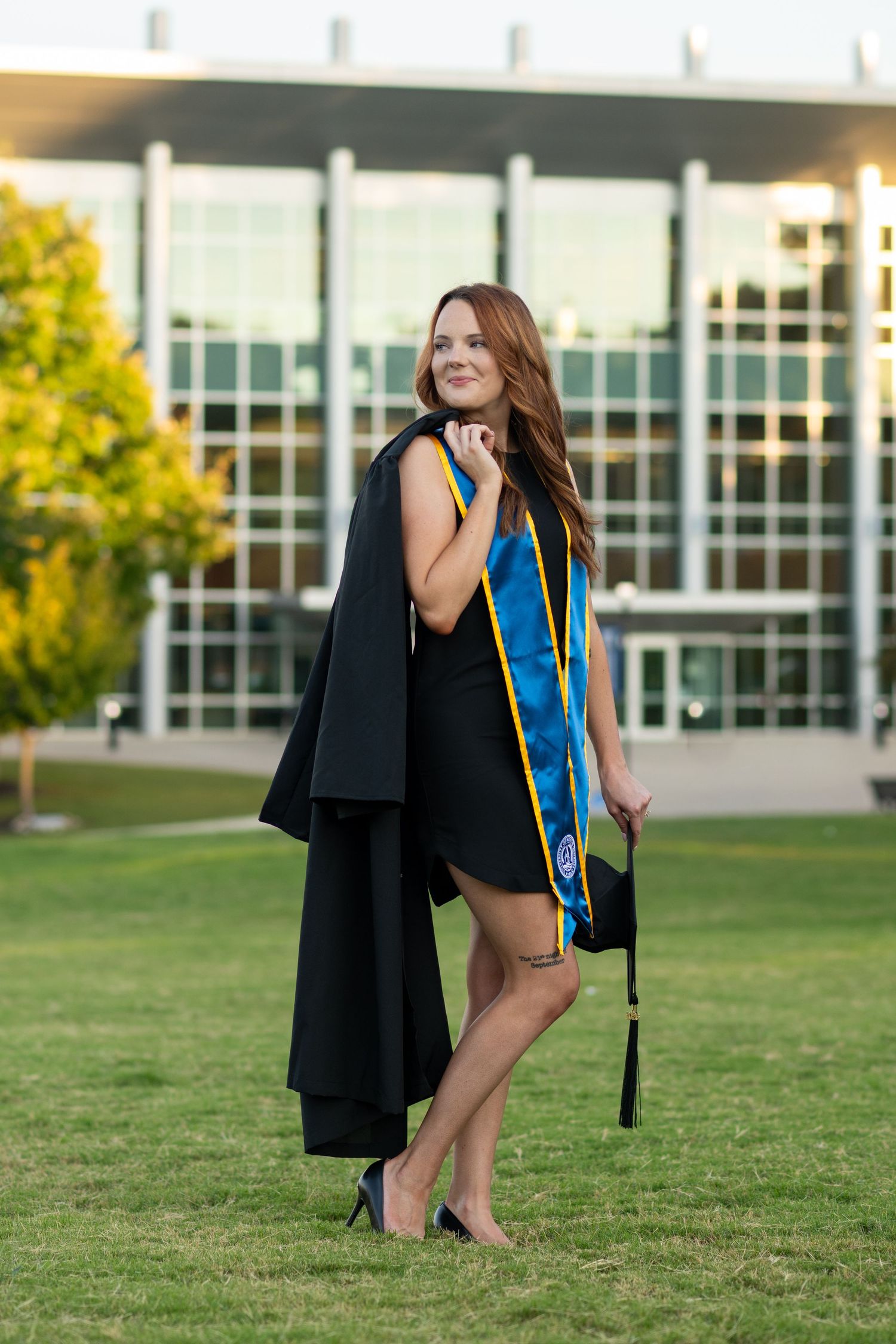 Young woman in graduation gown and blue stole holding cap on green campus lawn.