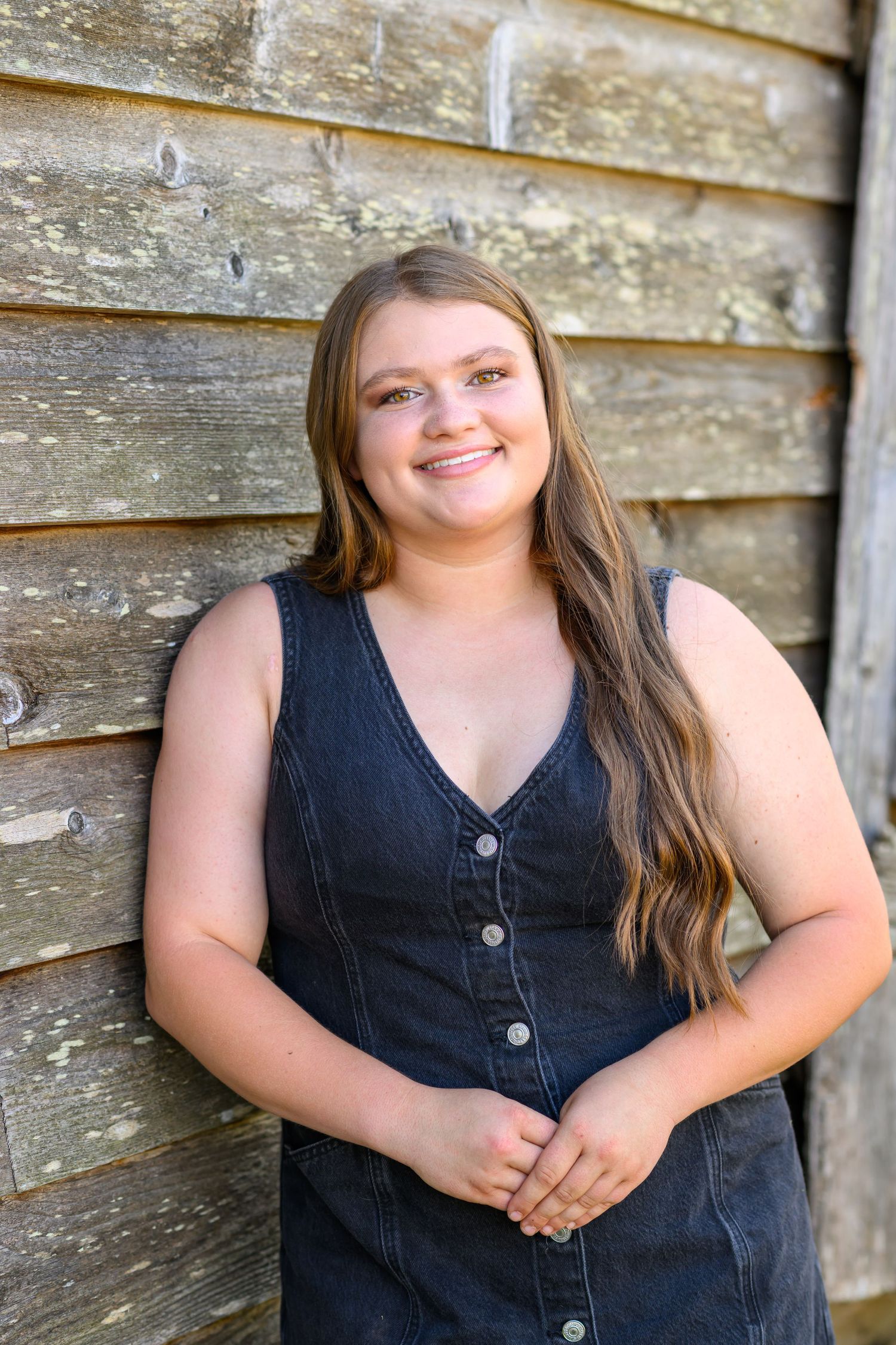 Young woman with long brown hair smiling in a dark denim dress, leaning against a rustic wooden wall outdoors.