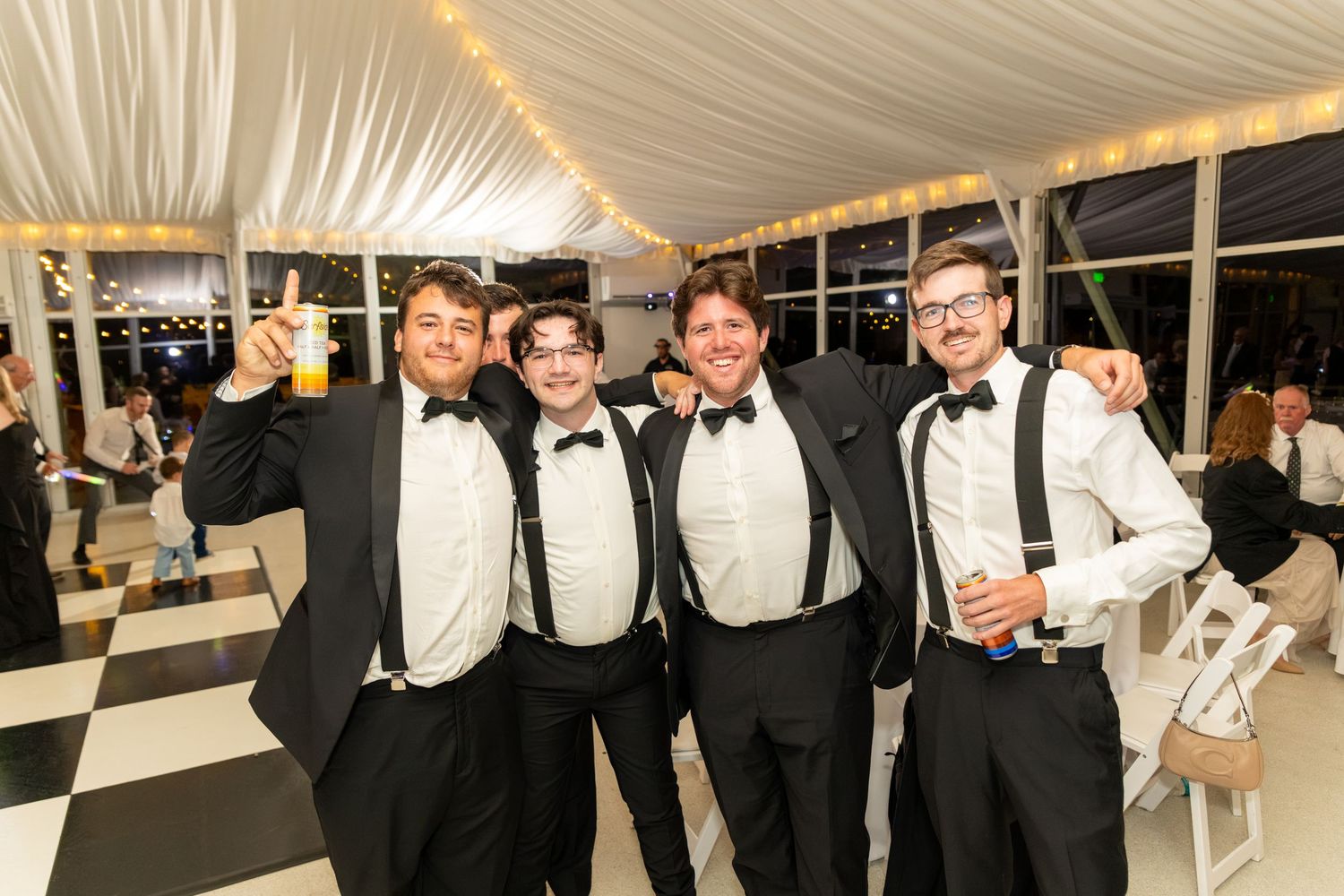 Four men in black tuxedos with bow ties smile and pose together at a formal event on a checkered dance floor.