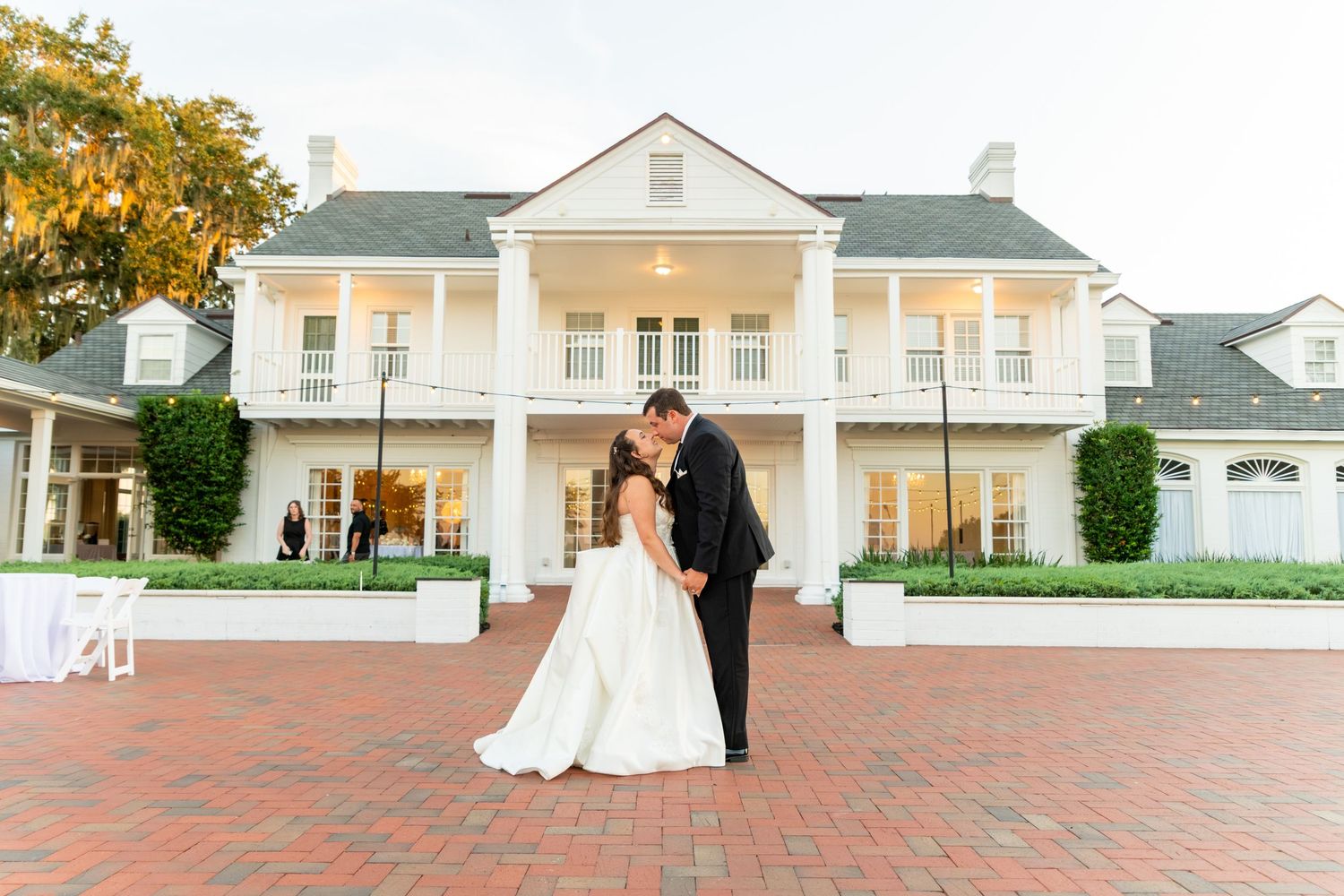 Bride and groom share romantic kiss in front of elegant white Southern mansion on brick courtyard at dusk.