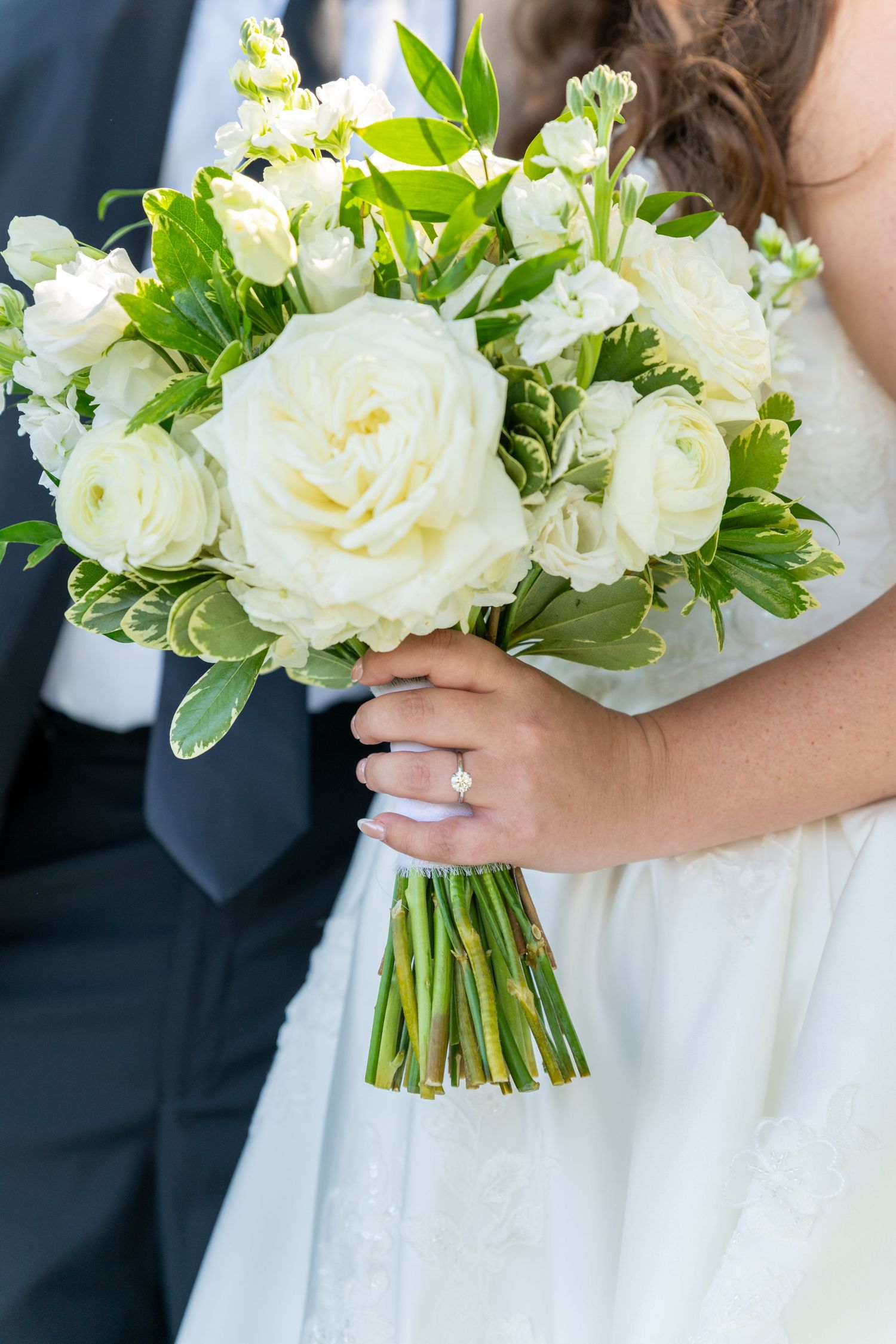 Bride holding a lush white rose and greenery wedding bouquet with a diamond engagement ring visible.