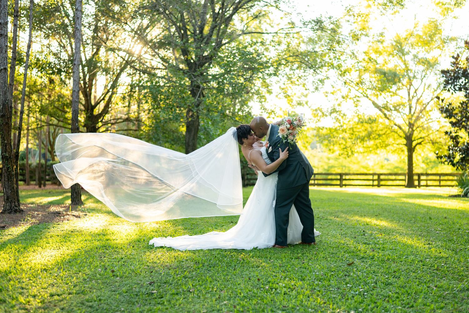 Bride and groom share romantic dip kiss outdoors, wedding veil flowing in sunlit garden.