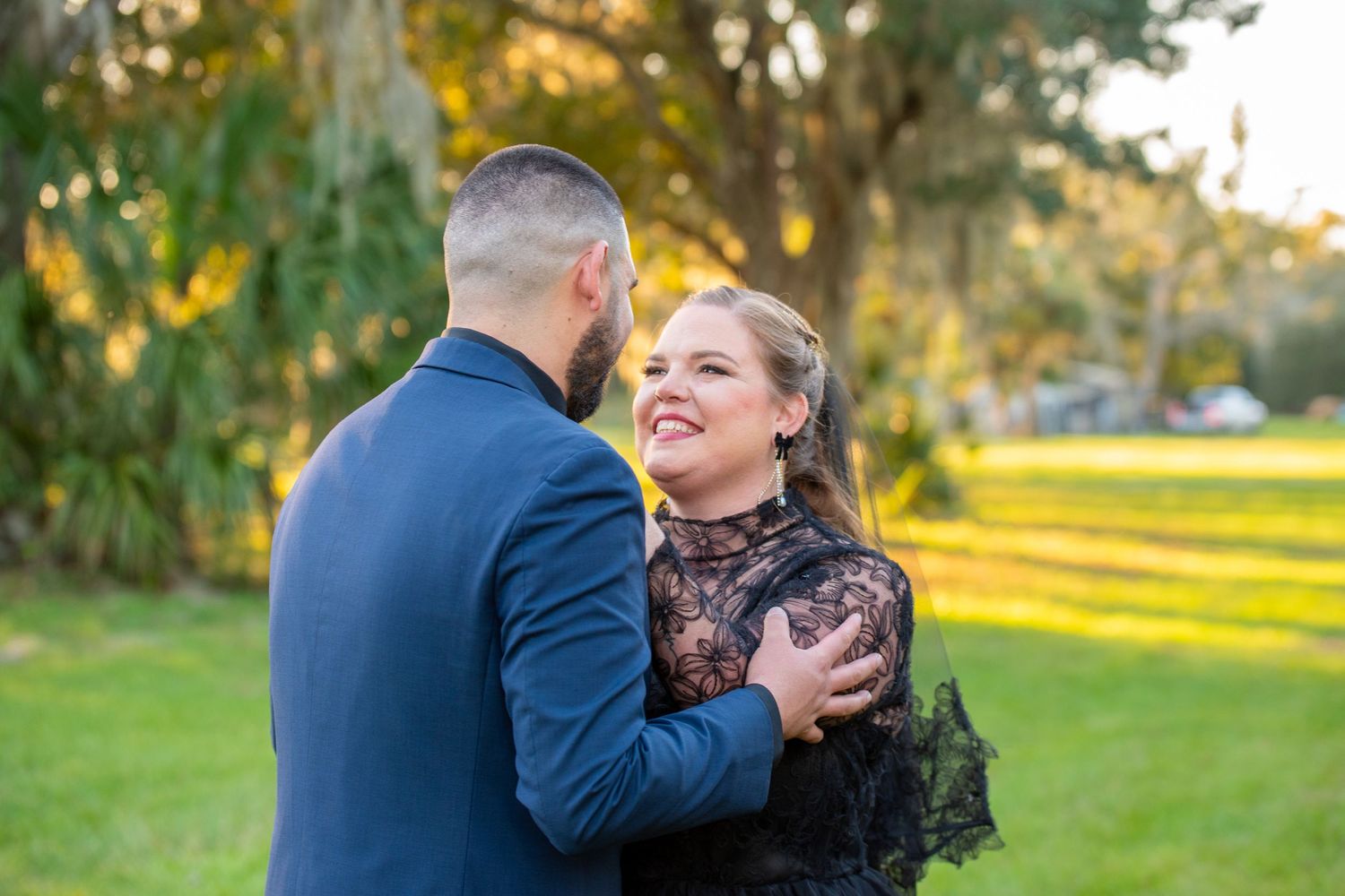 Couple in black lace and blue suit embracing outdoors during golden hour in a lush green park setting.