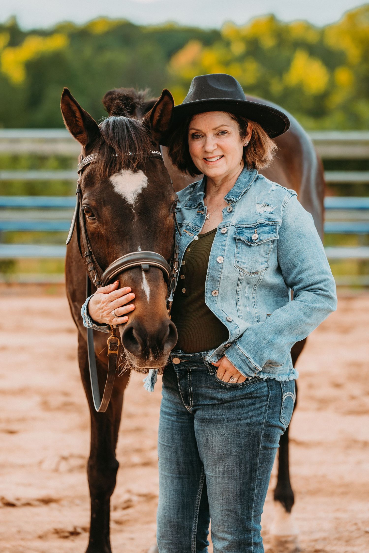 Woman in cowboy hat and denim jacket smiling beside a dark brown horse with white star marking in a sandy arena.