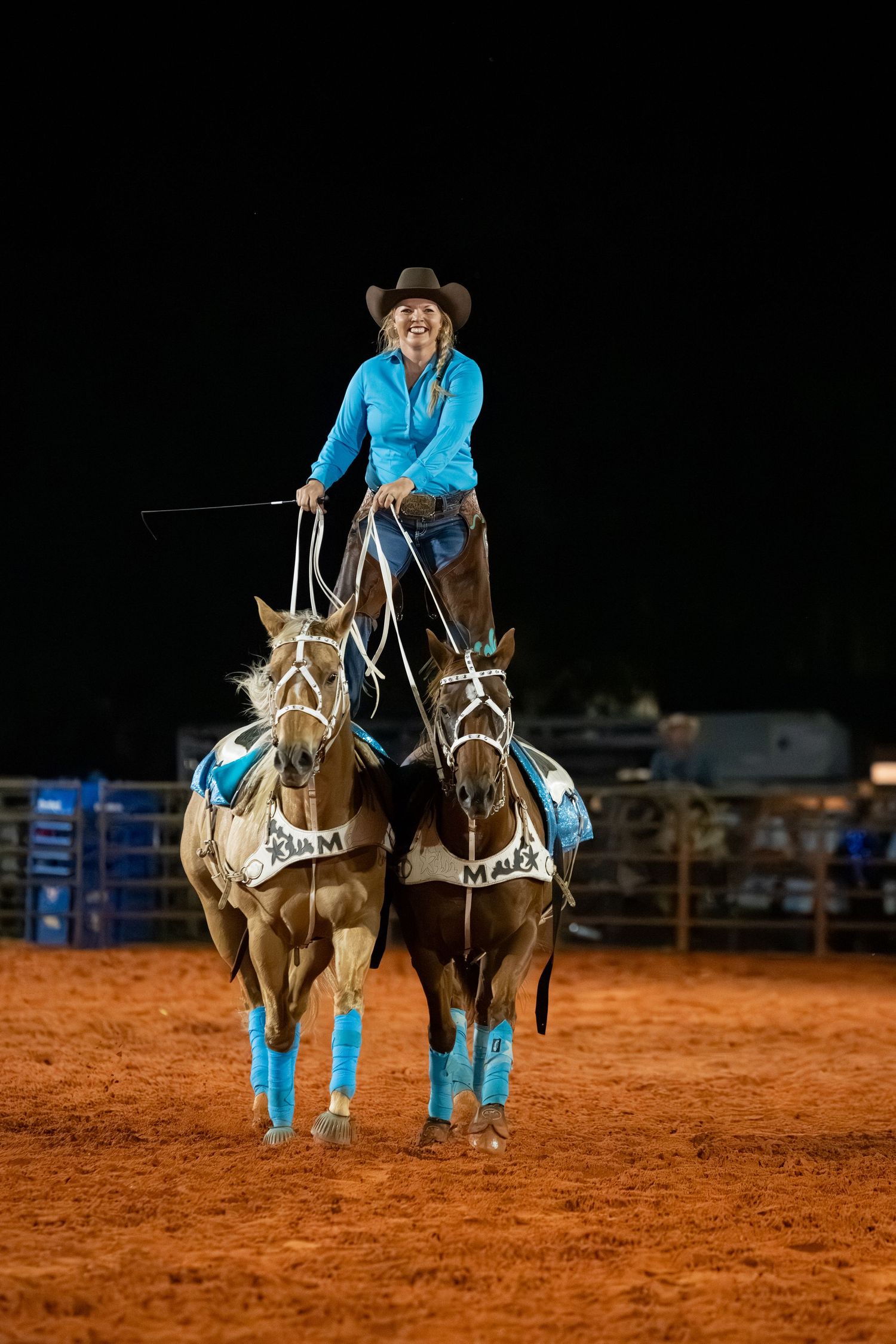 Smiling cowgirl in turquoise shirt stands atop two horses at a nighttime rodeo arena performance.