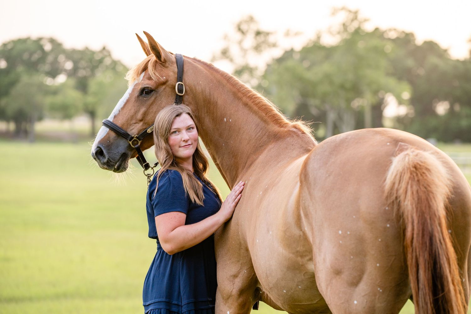 Woman in navy shirt poses with chestnut horse in sunny green pasture.