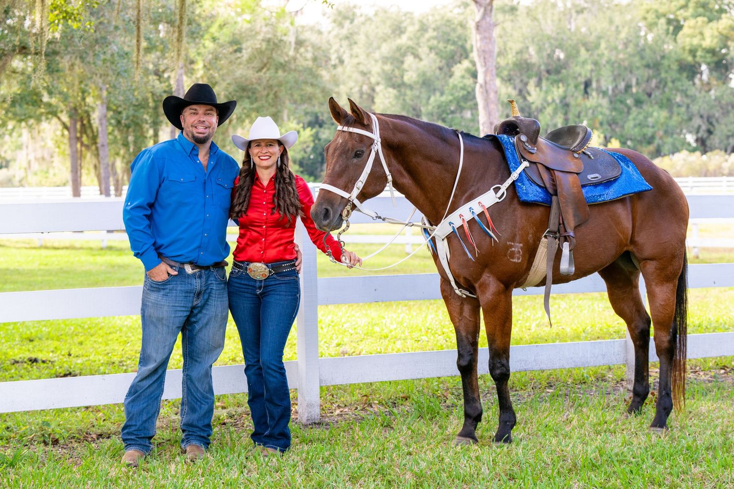 Couple in western attire standing with saddled chestnut horse by white fence outdoors.