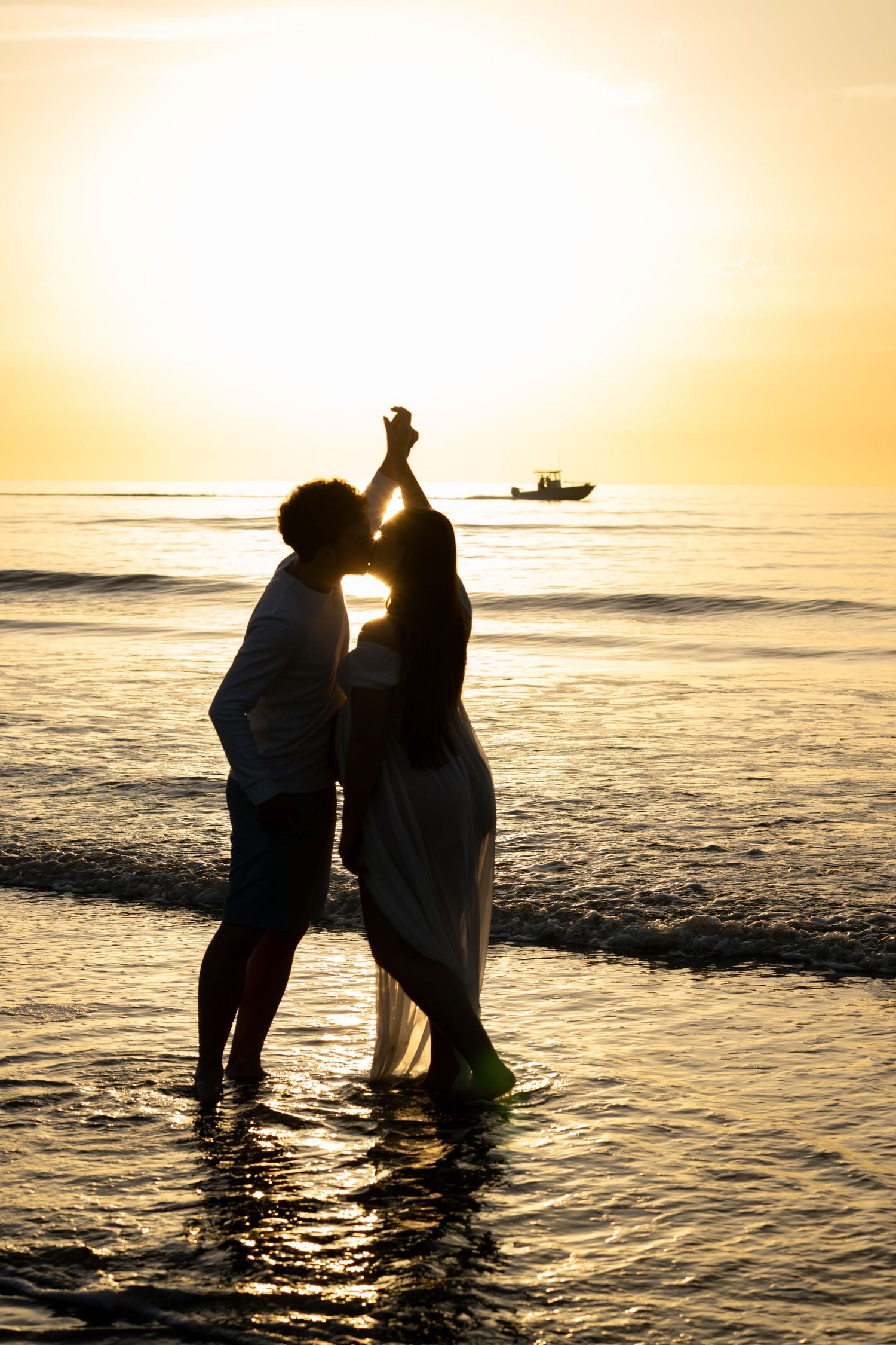 Romantic couple silhouetted against golden sunset on beach, dancing in ocean waves with boat in background.