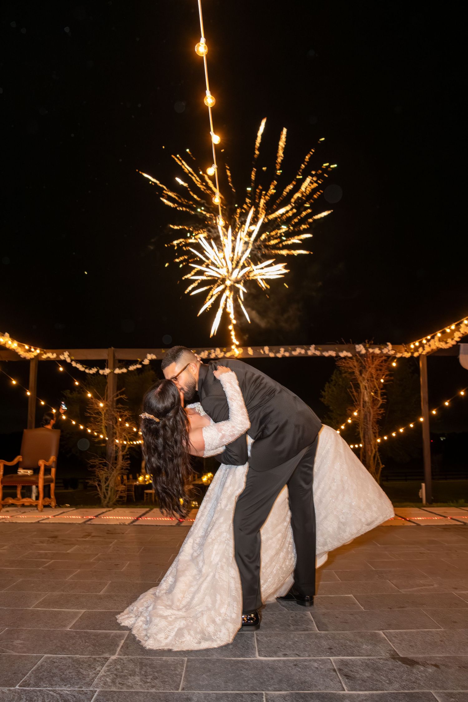 Bride and groom share a romantic dip kiss with fireworks bursting in the night sky above.