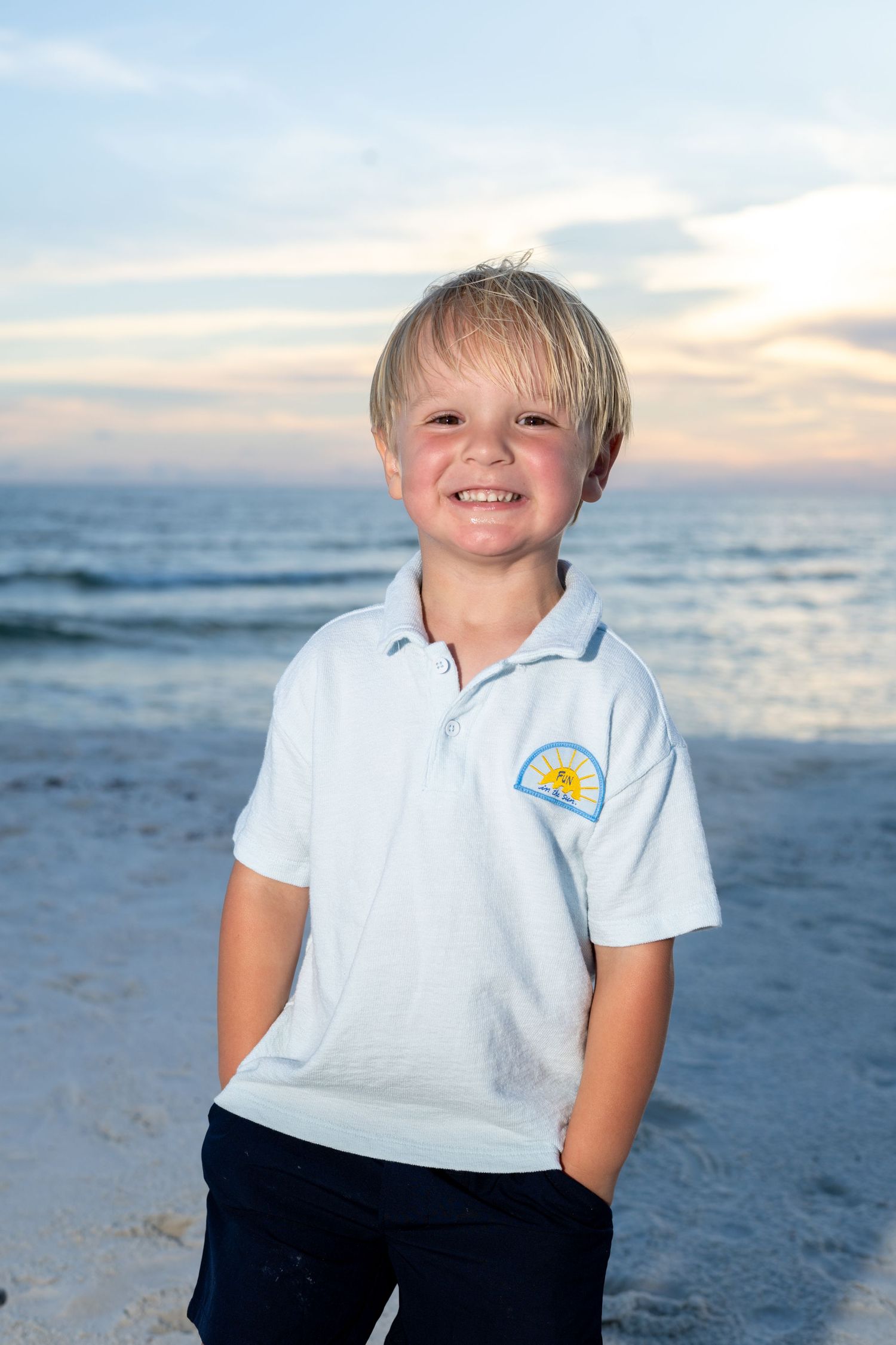 Smiling blonde boy in a white polo shirt posing on a beach at sunset.