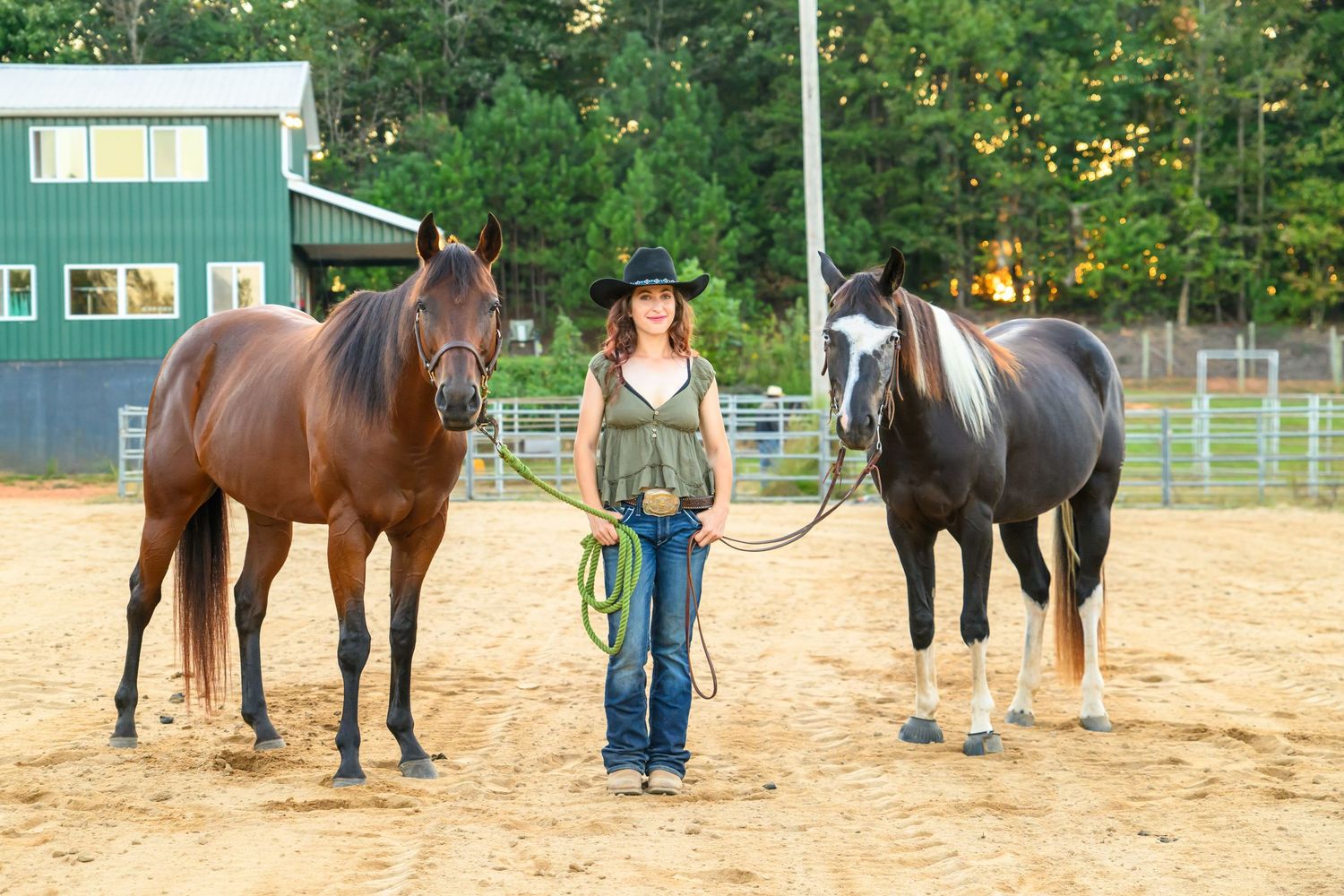 Woman in cowboy hat holding leads for two horses in sandy arena with green barn background.
