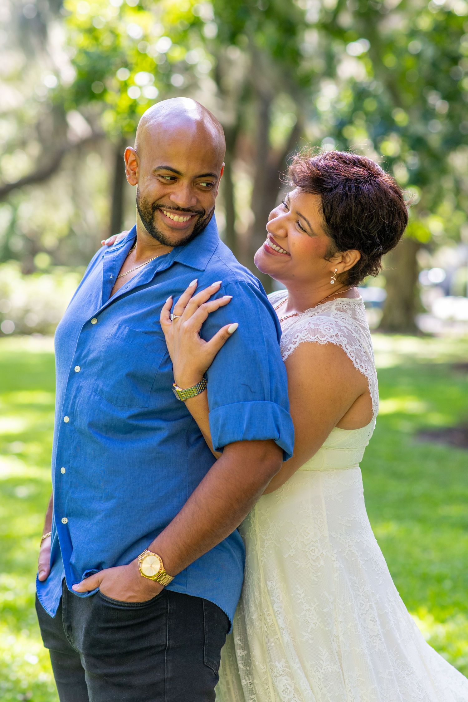 Happy couple smiling and embracing outdoors in a lush green park, man in blue shirt, woman in white dress.