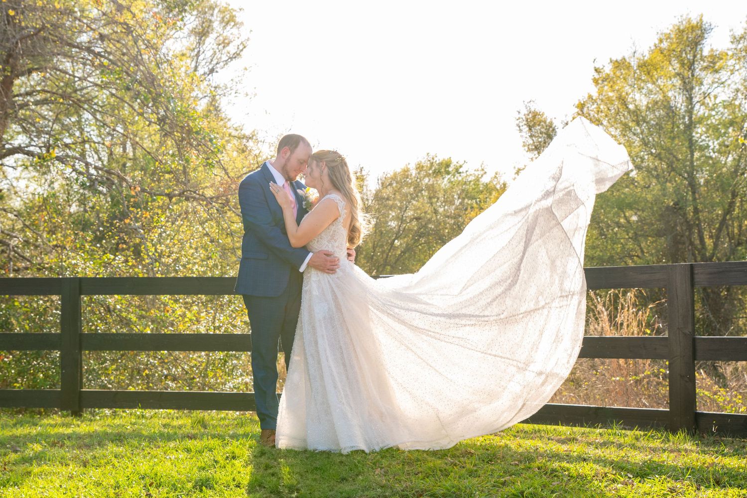 Bride and groom embrace at golden hour as her gown flows in the breeze beside a wooden fence.