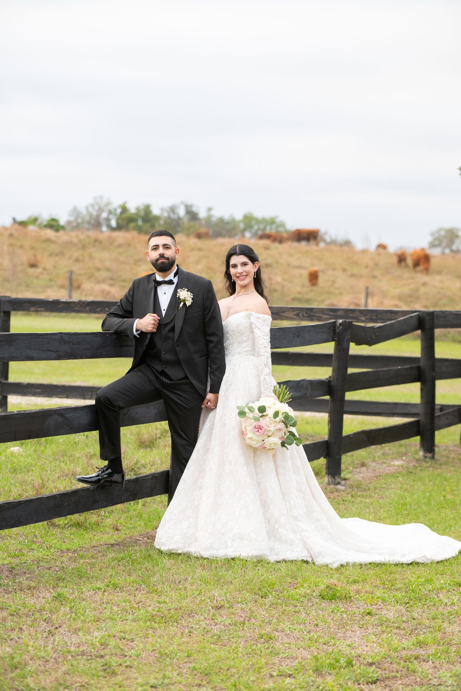 Bride and groom pose by a black wooden fence with cattle grazing in the background at a farm wedding.