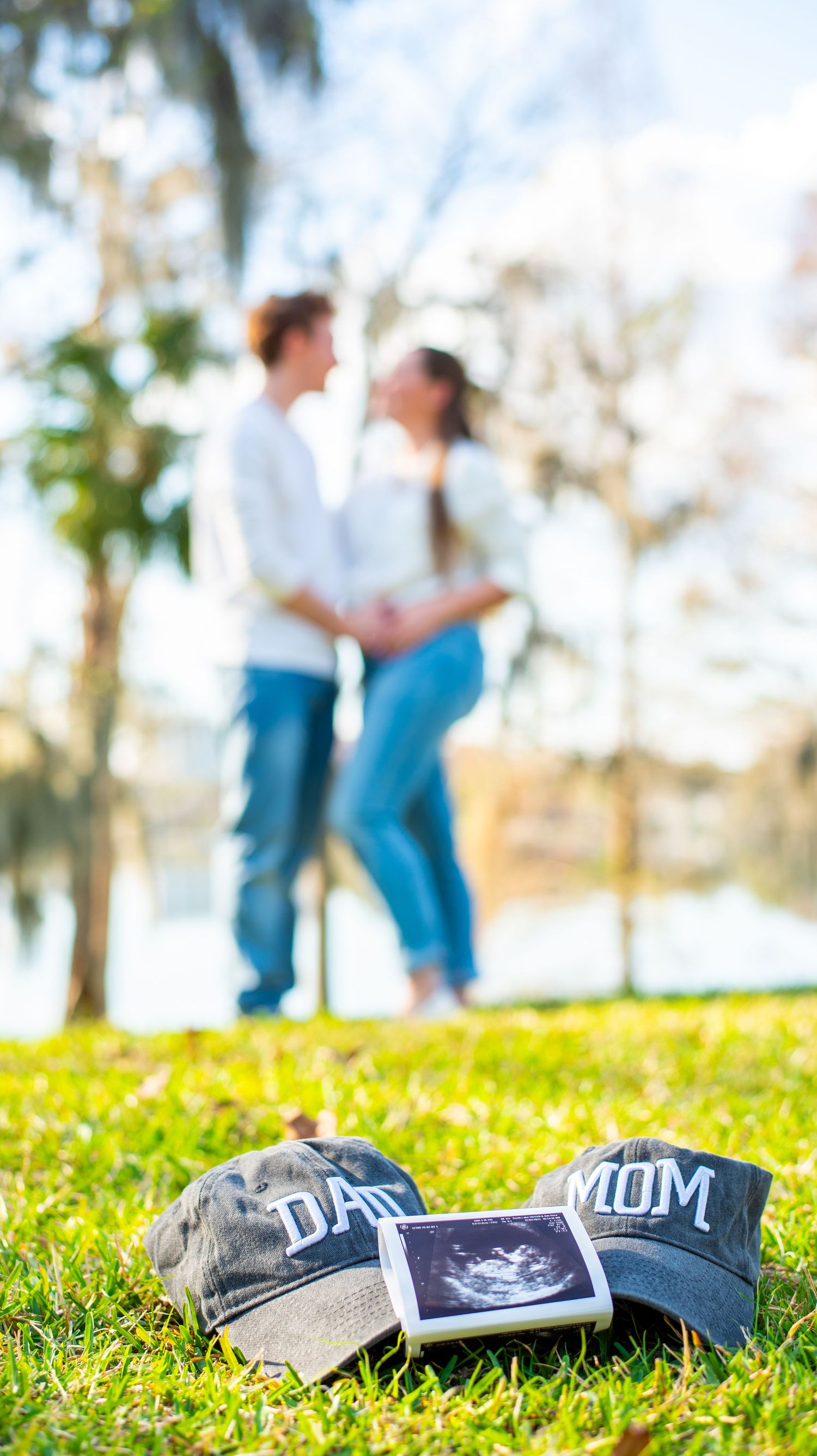 Dad and Mom hats with ultrasound photo on grass as couple embraces in the background.