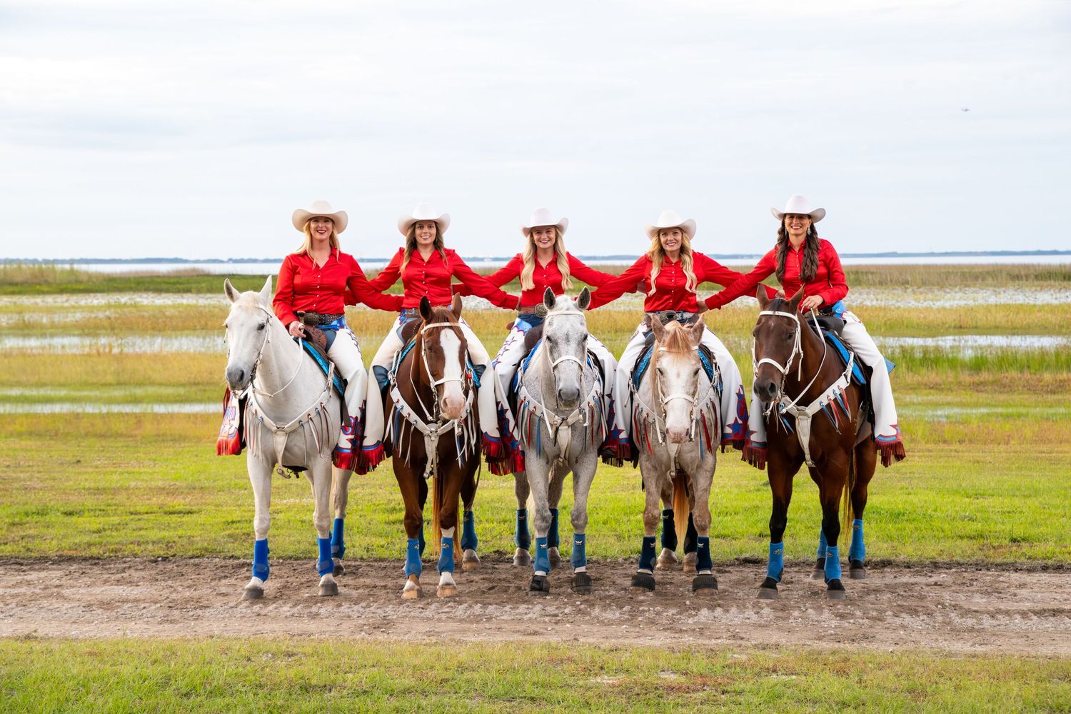 Five women in red shirts and white cowboy hats pose on horseback in a scenic outdoor setting.