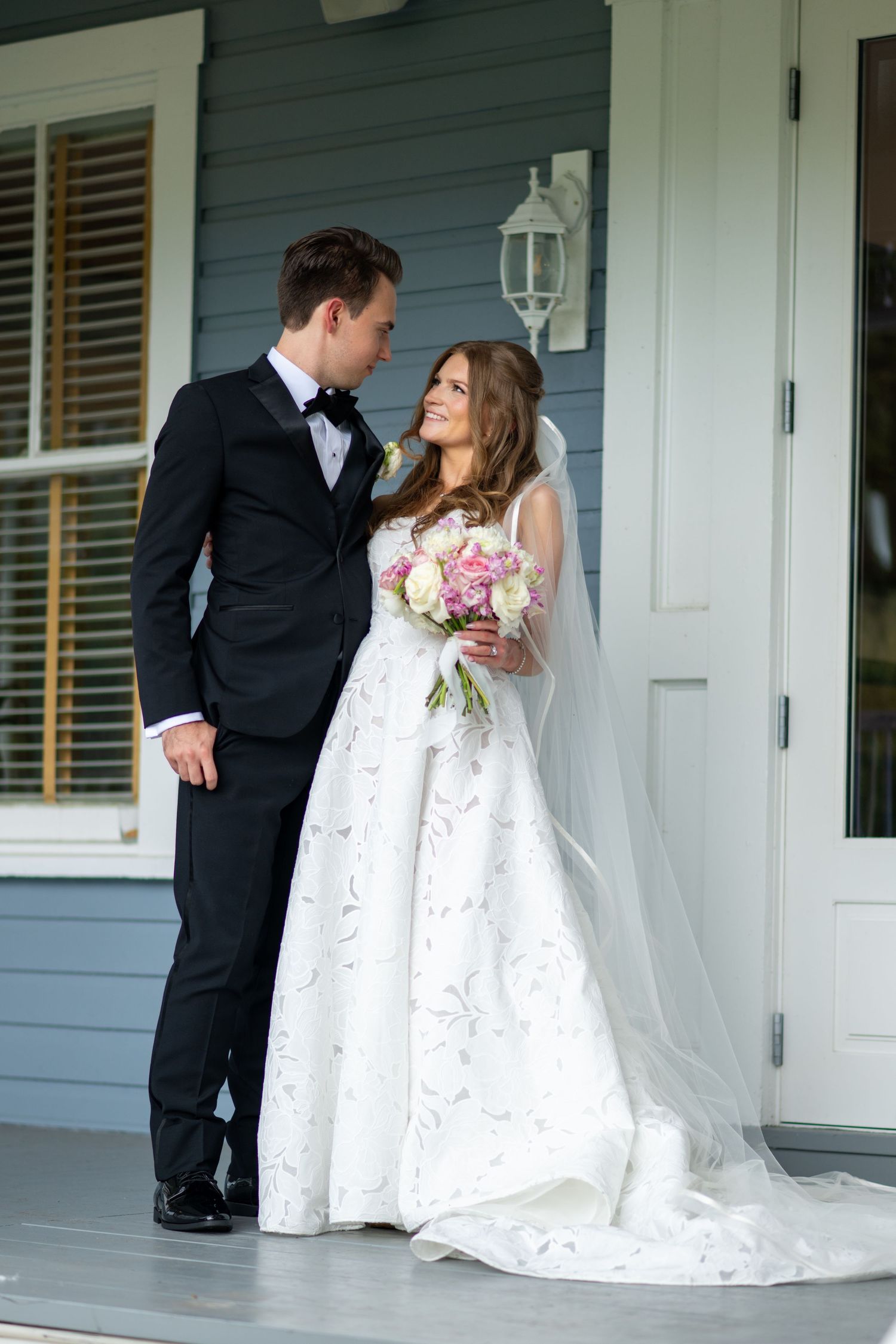 Bride and groom sharing romantic moment on porch, bride holding pink and white bouquet.