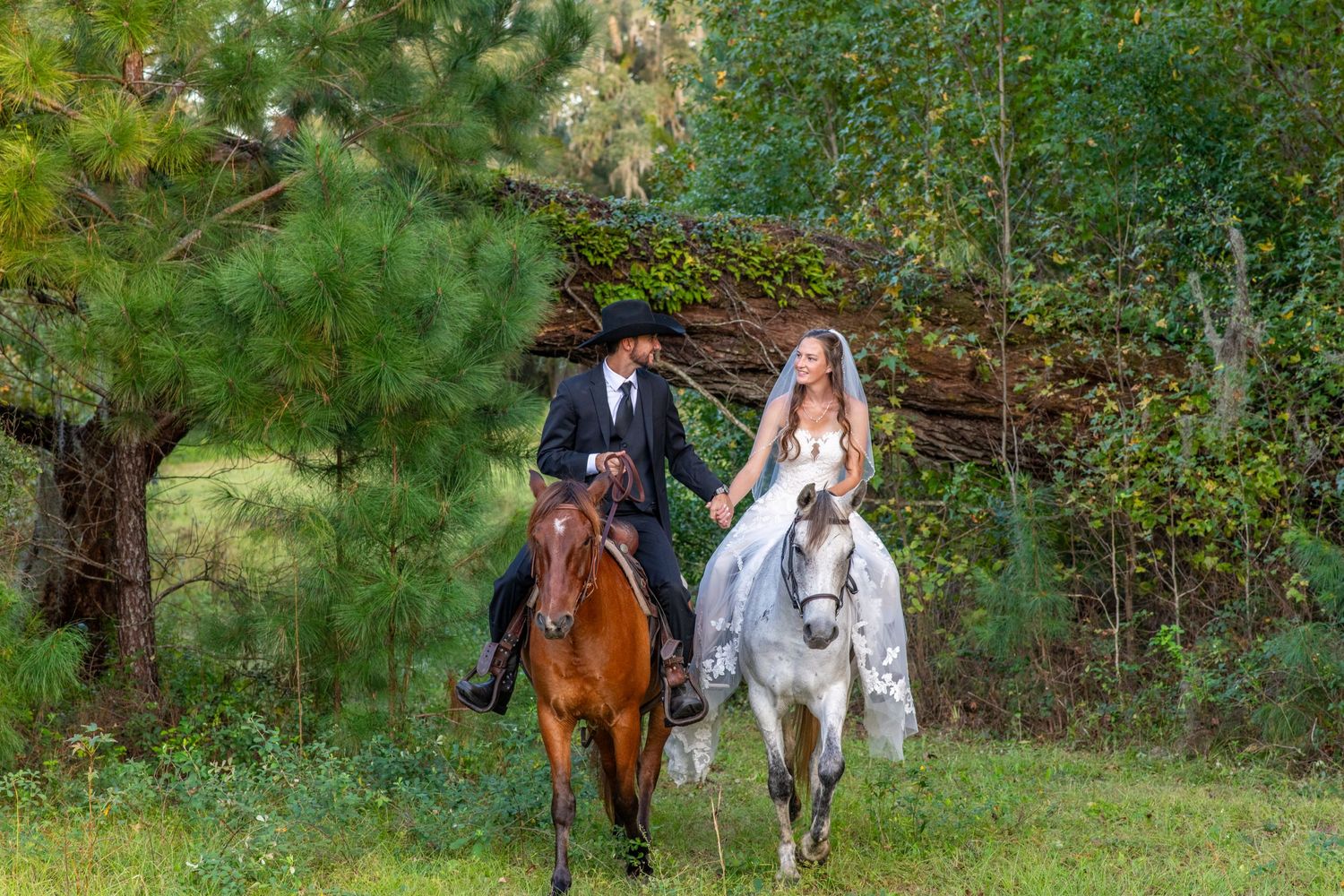 Bride and groom ride horses through a lush green forest in a romantic outdoor wedding portrait.
