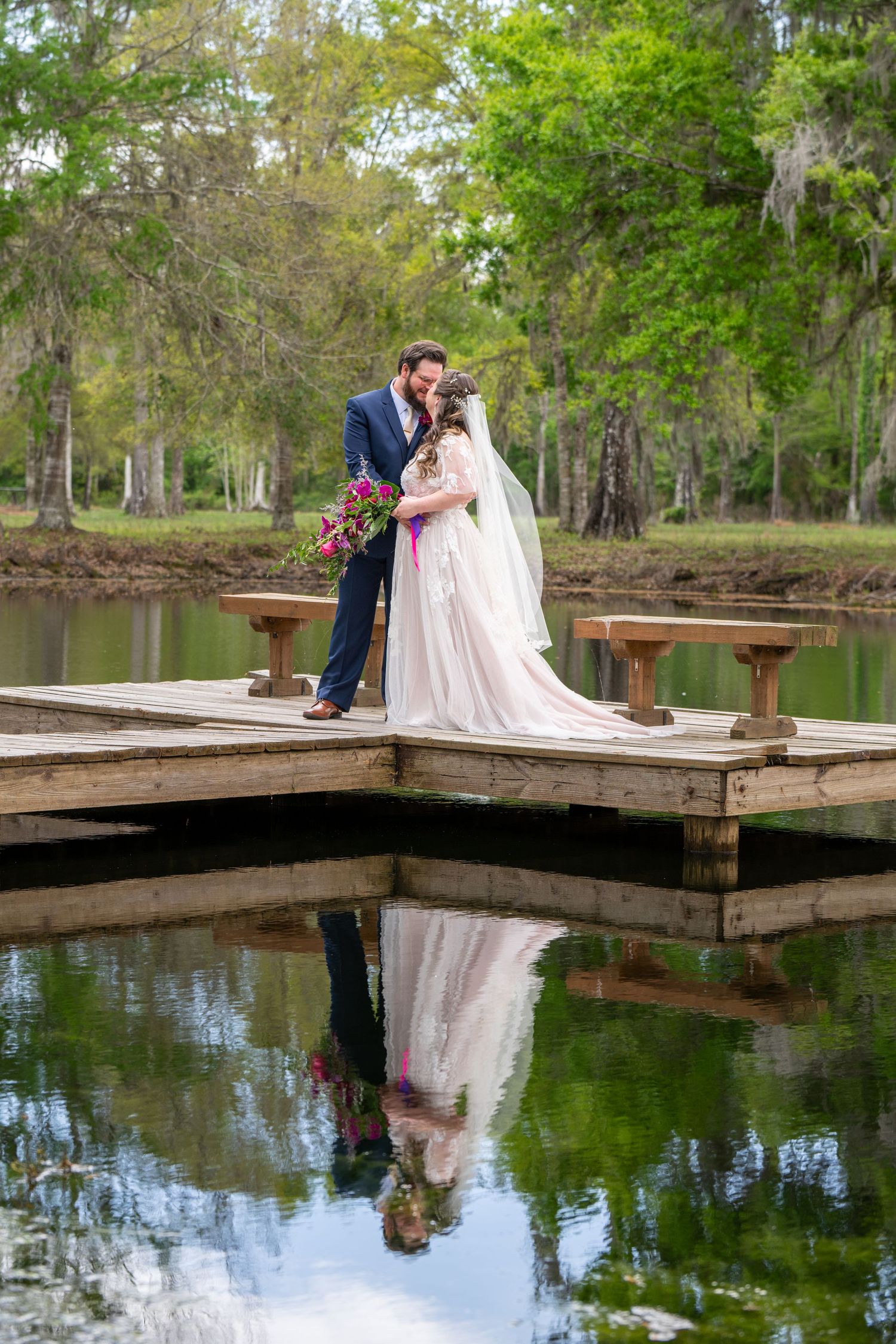 Bride and groom kiss on a wooden dock with their reflection mirrored in the still lake below.
