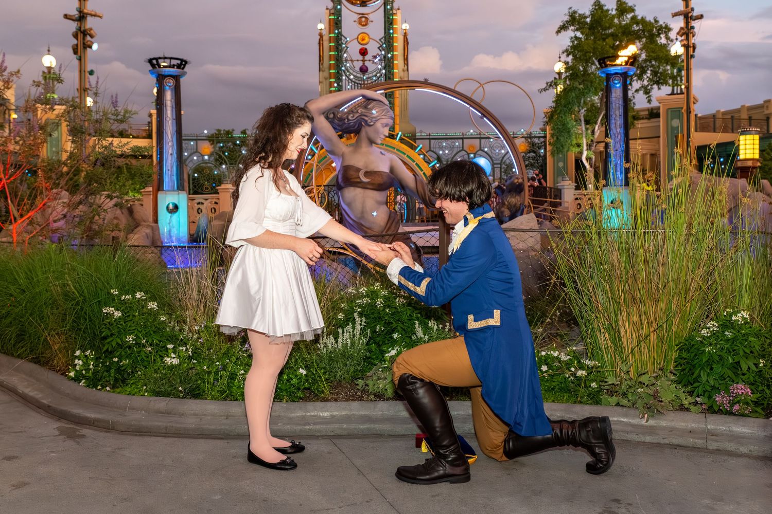 A man in a blue costume proposes on one knee to a woman in a white dress in a garden at dusk.