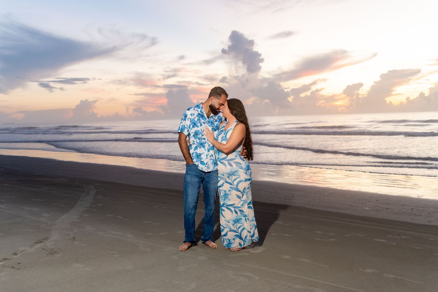 Couple embracing on beach at sunset, man in floral shirt and woman in blue floral dress, ocean waves behind them.
