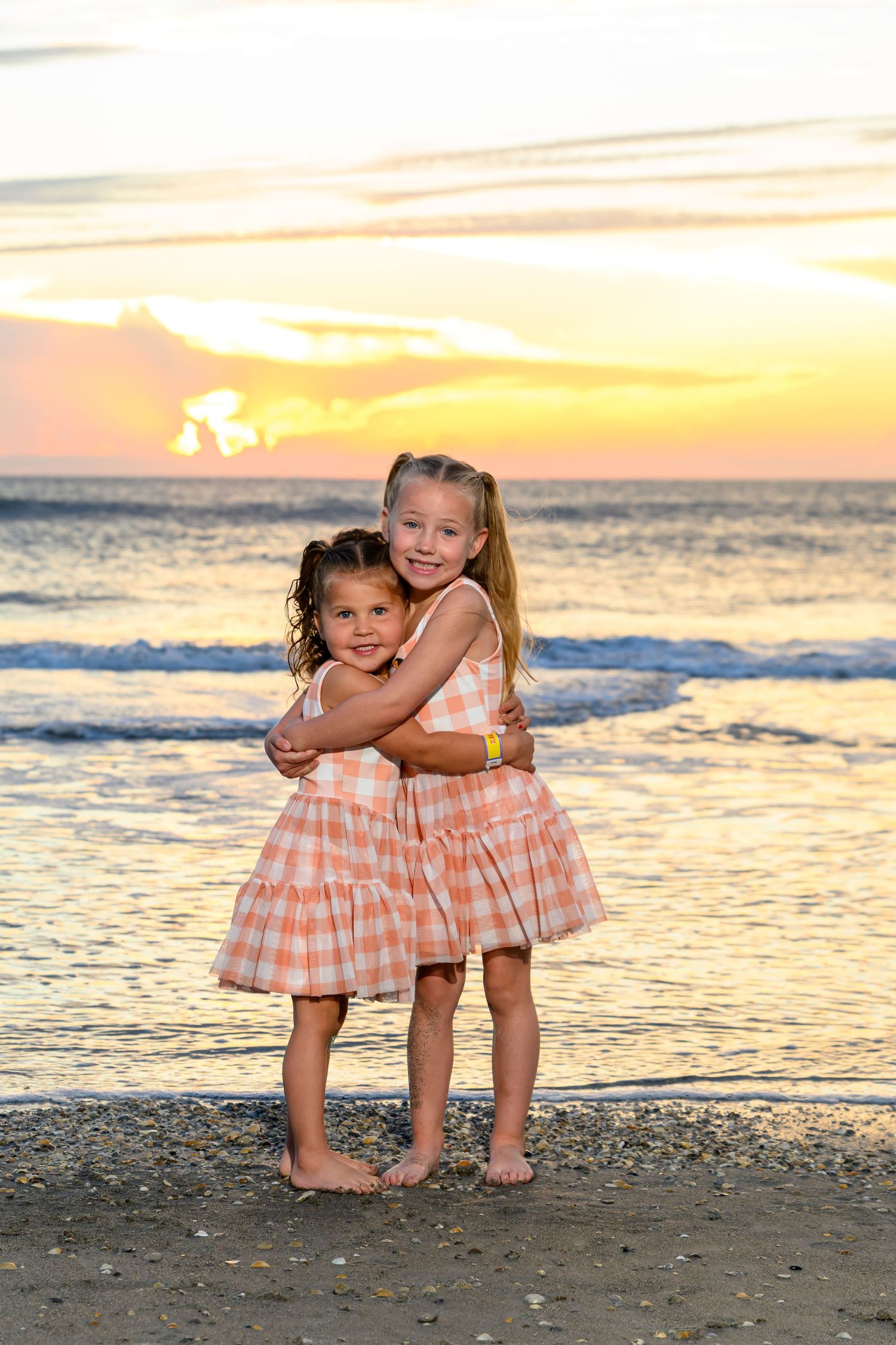Two young girls in matching peach plaid dresses hug on a sandy beach at sunset with gentle waves and a glowing orange sky behind them.